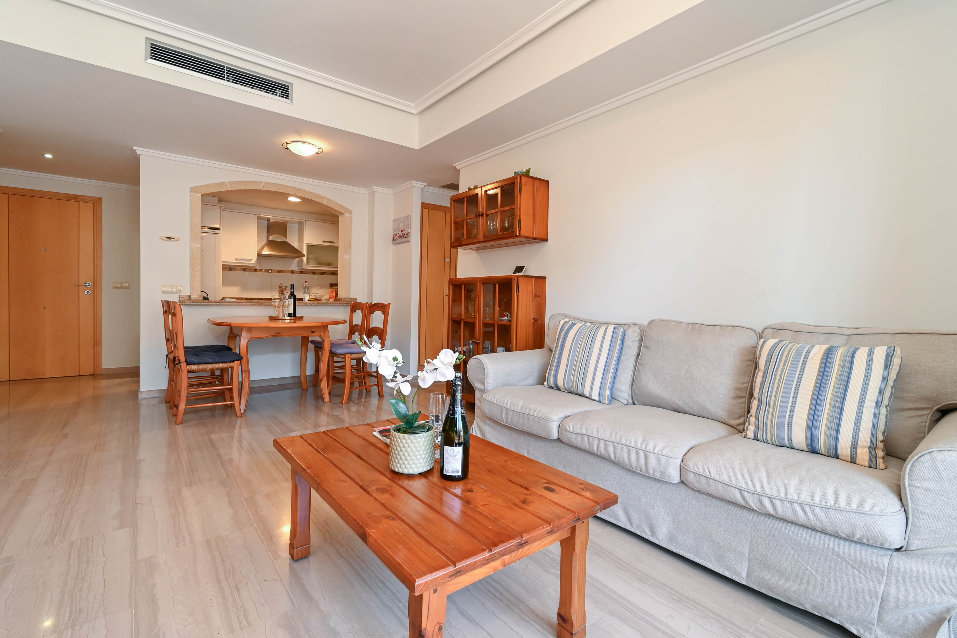 Salón comedor de planta abierta con suelos de mármol, sofá beige, mobiliario de madera maciza y un arco de piedra que conecta con la cocina equipada.