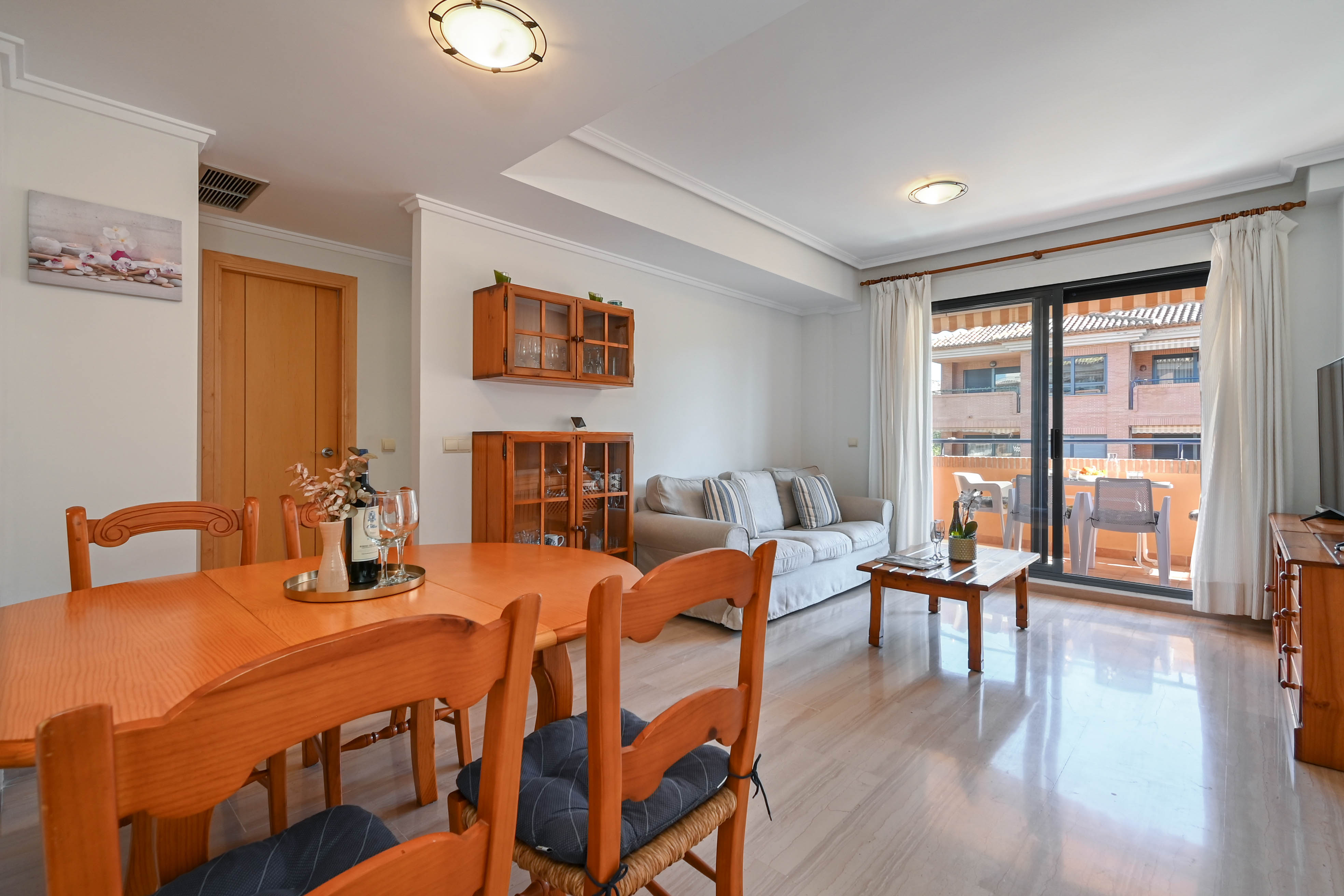 Salón comedor luminoso con mobiliario de madera, suelos de mármol pulido y acceso directo a una terraza amueblada a través de grandes ventanales corredizos.
