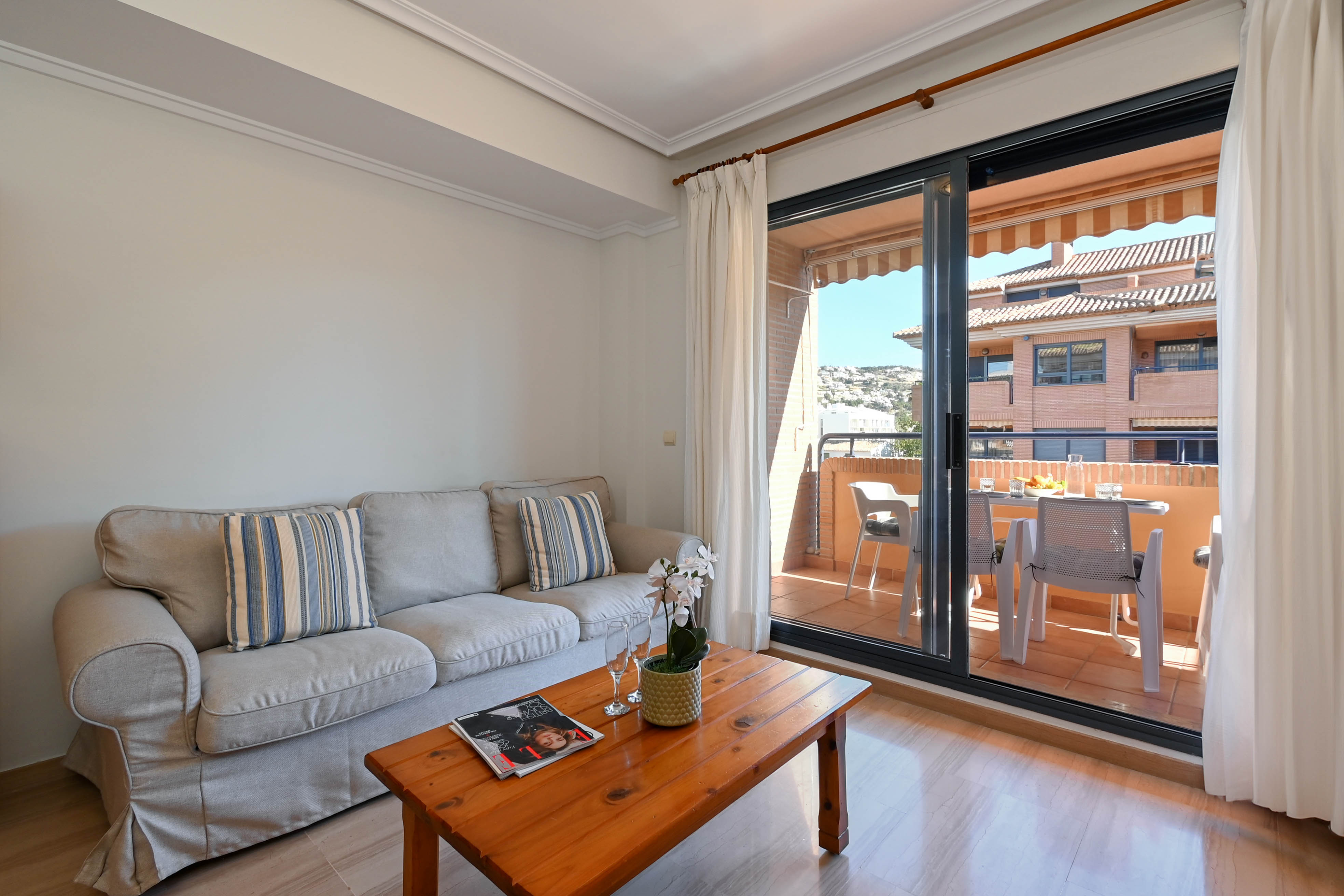 Salón luminoso con sofá gris y mesa de madera, conectado mediante una puerta corredera a una terraza amueblada con vistas a la zona residencial.