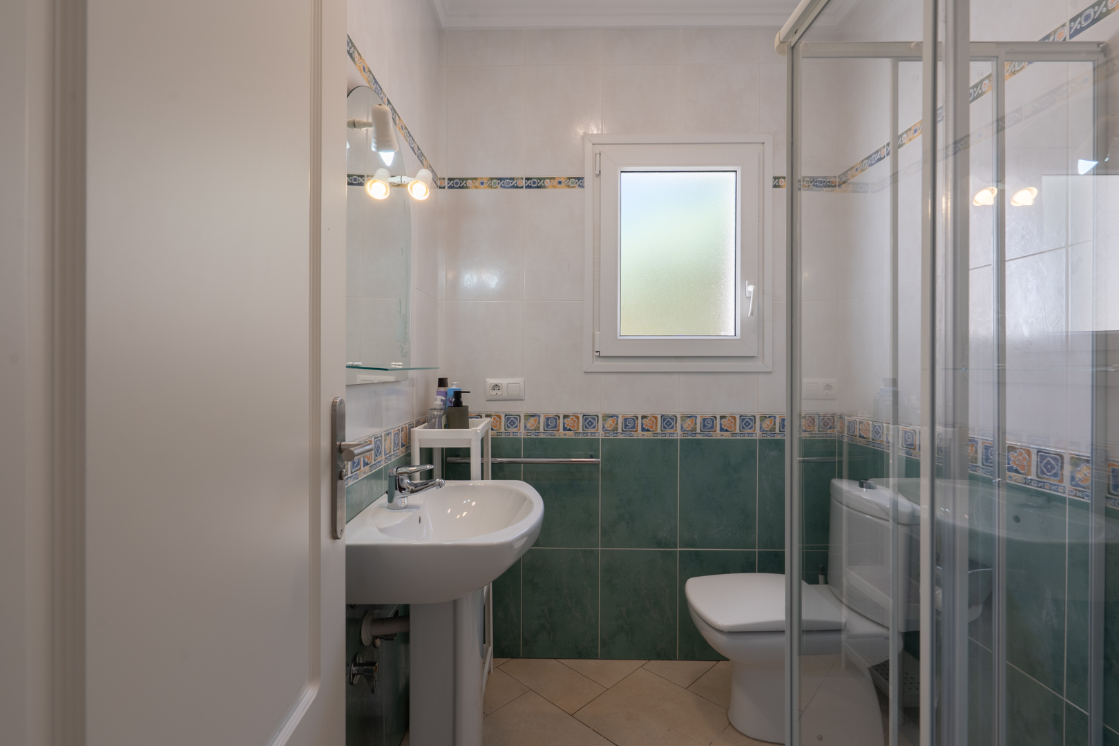 Baño funcional con azulejos verdes y blancos, lavabo de pedestal, ducha con mampara de cristal y ventana de PVC con cristal traslúcido.