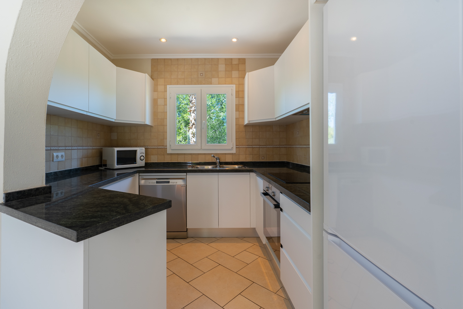 Cocina funcional en forma de U con encimeras de granito oscuro, mobiliario blanco, electrodomésticos integrados y una ventana de PVC con vistas al exterior.