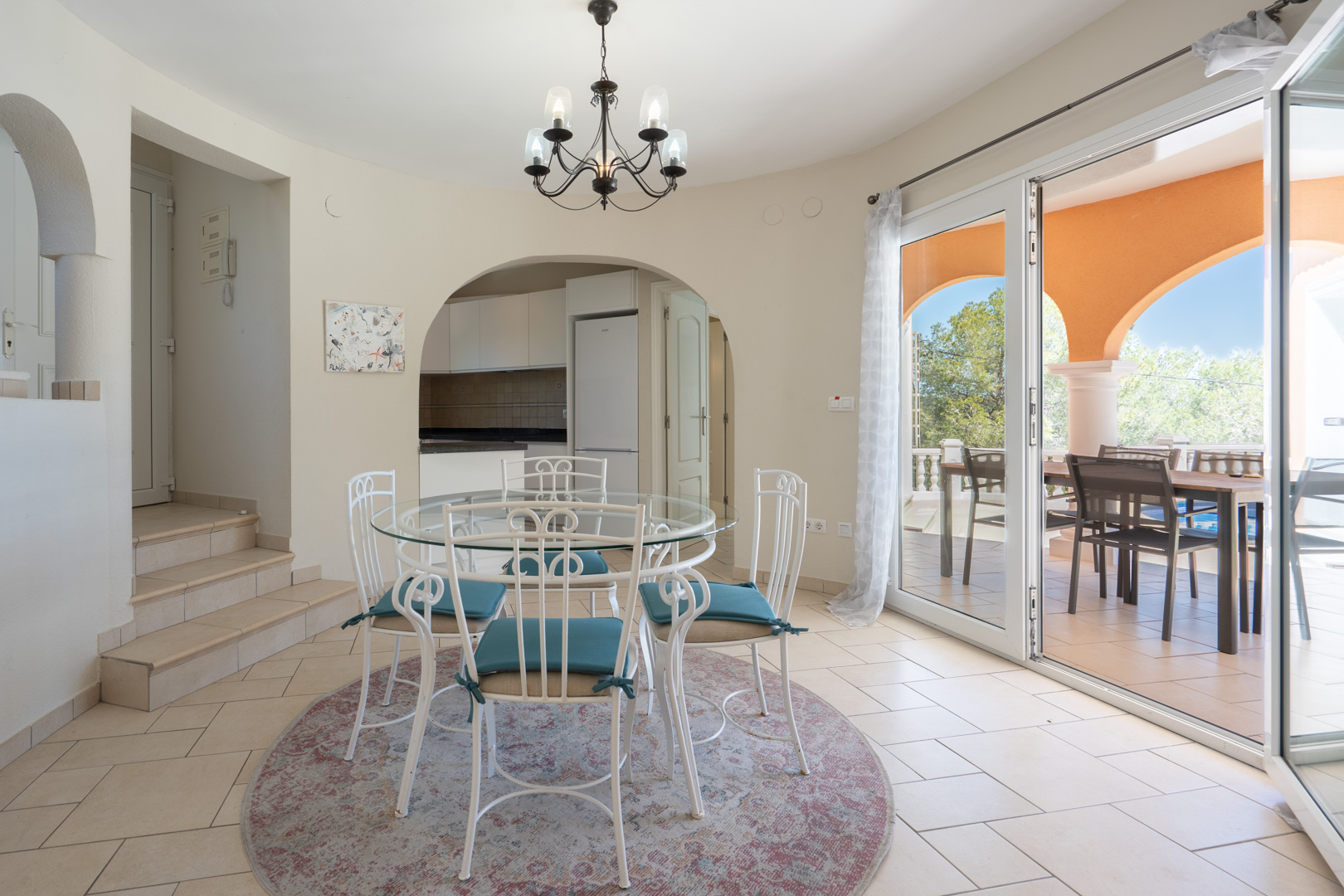 Comedor luminoso con mesa de cristal y sillas de forja blanca, conectado a la cocina mediante un arco y con acceso directo a la terraza cubierta.