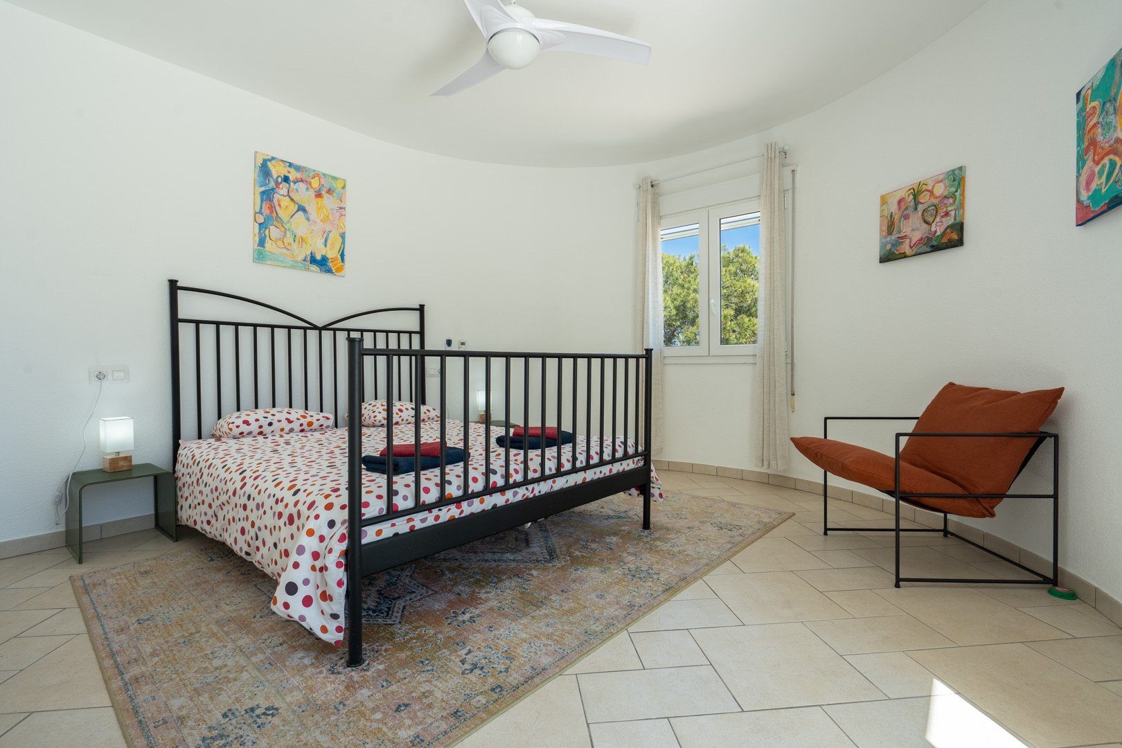 Dormitorio luminoso con pared curva, cama de forja negra, suelos de baldosa clara y ventana de PVC con vistas al jardín mediterráneo.