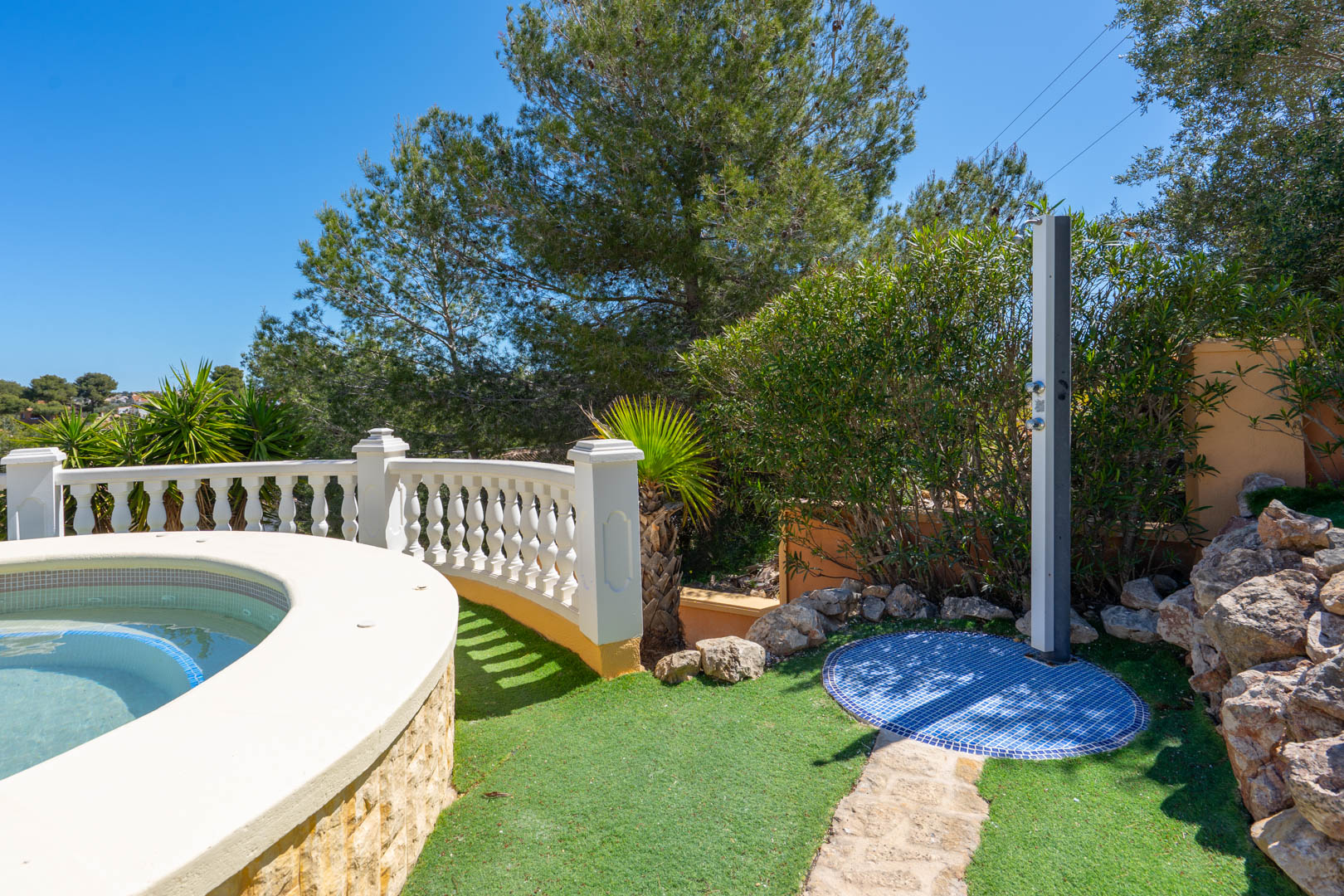 Ducha exterior con base de mosaico azul junto a la piscina de borde curvo, rodeada de vegetación autóctona y una balaustrada blanca clásica.