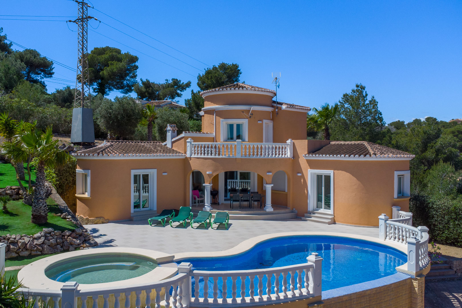 Exterior de la villa con piscina de diseño curvo, jacuzzi integrado y amplia terraza pavimentada. La fachada mediterránea destaca por sus arcos y balaustradas blancas.