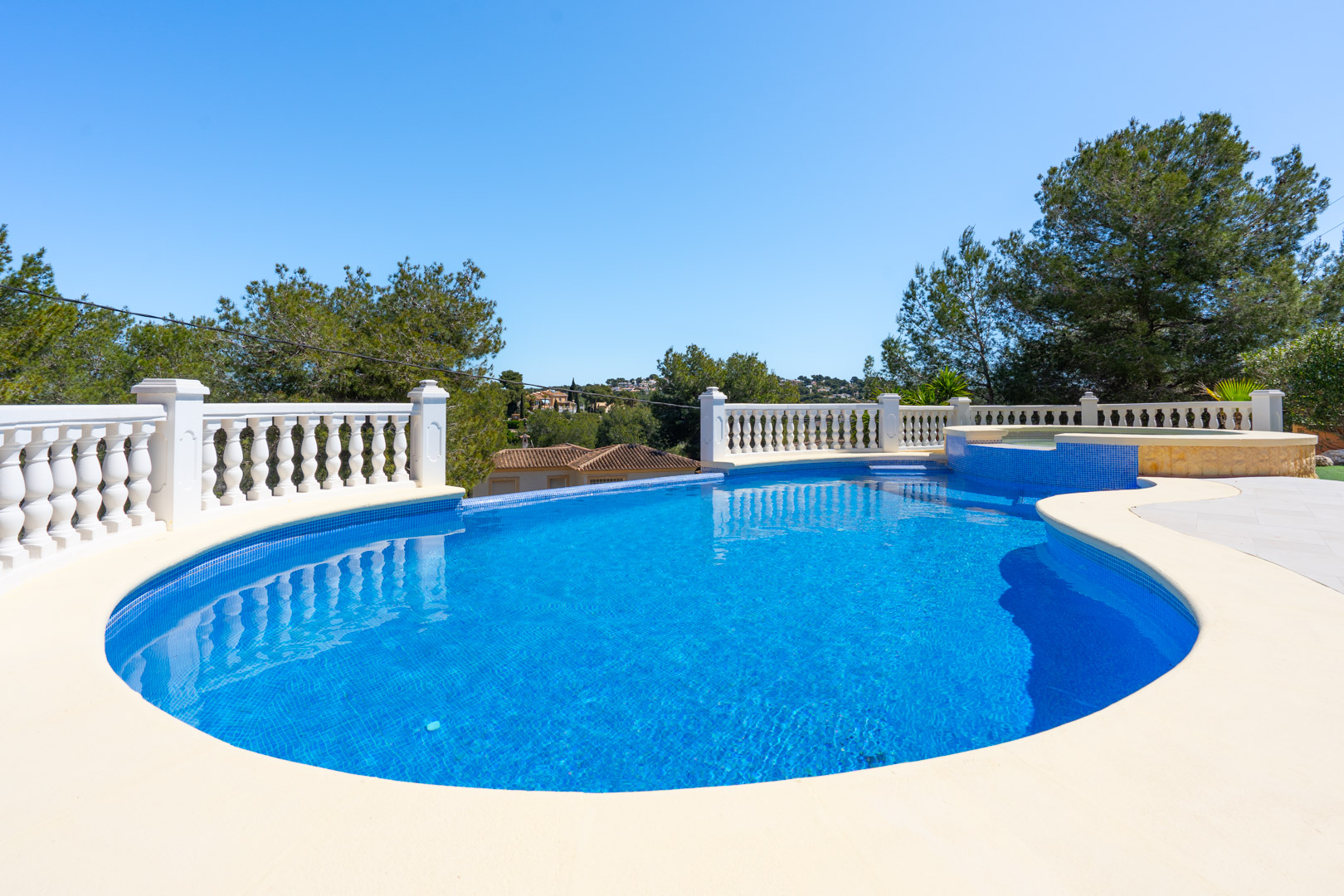 Piscina exterior de forma irregular con gresite azul, rodeada por una terraza pavimentada y una balaustrada blanca clásica con vistas al entorno arbolado.