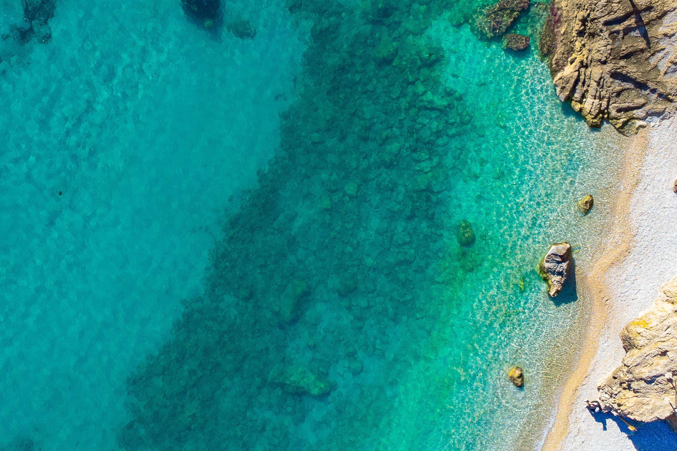 Vista aérea de la costa de Jávea, mostrando aguas cristalinas de color turquesa, un fondo marino rocoso y una orilla de guijarros naturales.