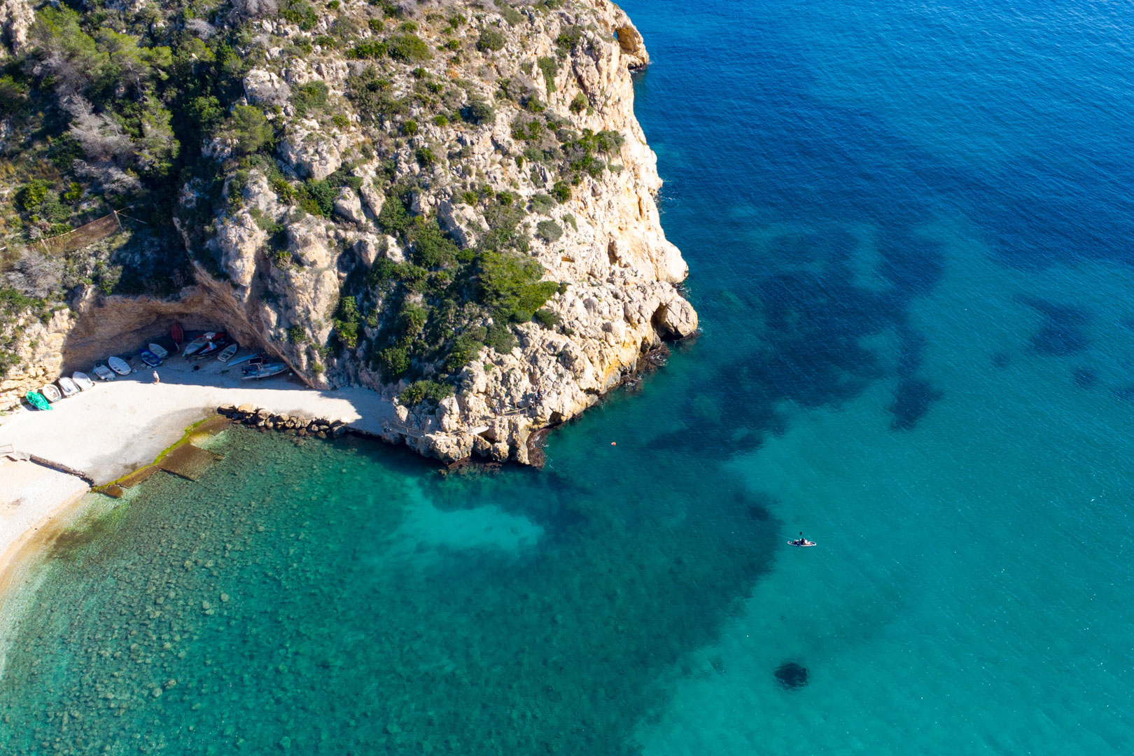 Vista aérea de una cala virgen en Jávea, con aguas turquesas cristalinas, acantilados escarpados y pequeñas embarcaciones resguardadas bajo un saliente de roca natural.