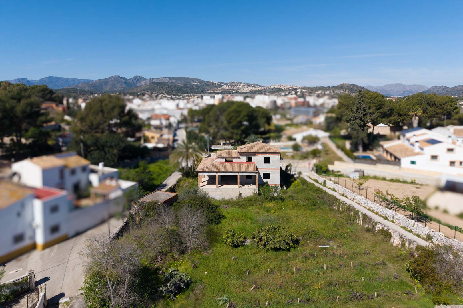 Vista aérea de la estructura de obra con tejado de teja cerámica sobre una amplia parcela de 1800 m², rodeada de vegetación y con vistas a las montañas.
