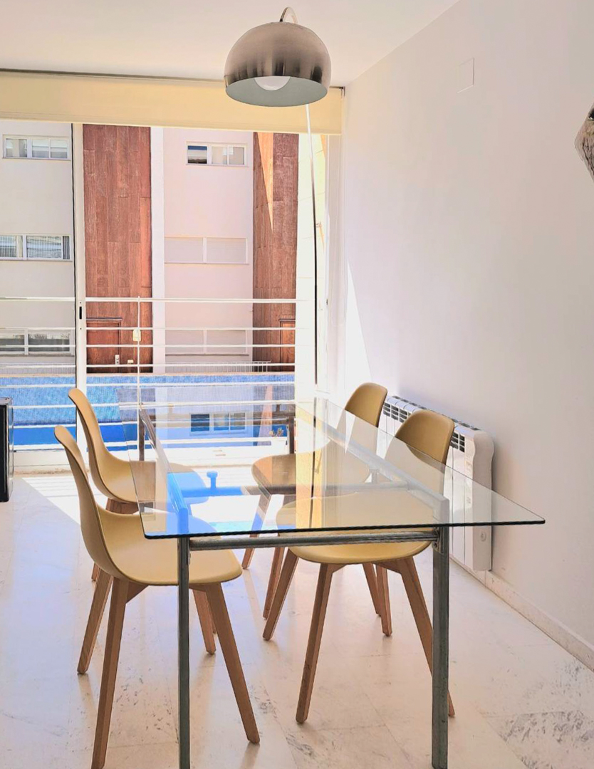 Comedor luminoso con mesa de cristal, sillas de estilo nórdico y suelo de mármol, con vistas directas a la piscina comunitaria a través del gran ventanal.