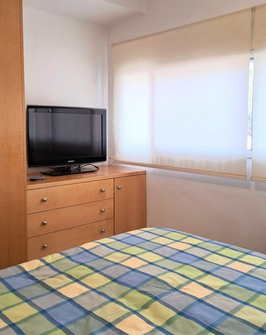 Dormitorio con mobiliario de madera a medida, televisor sobre cómoda y ventana con estores translúcidos que filtran la luz natural.