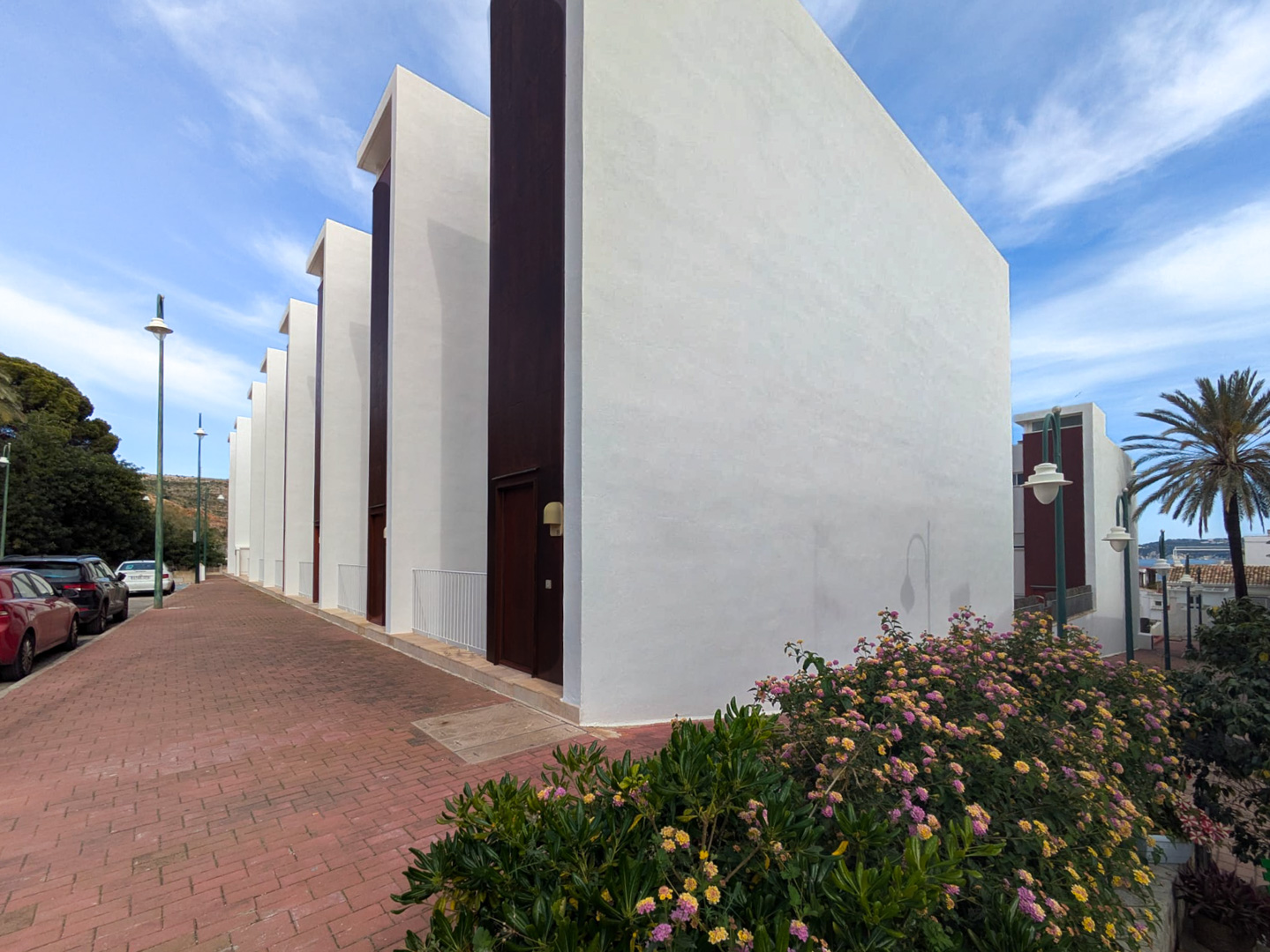 Fachada exterior de diseño contemporáneo con muros blancos y paneles verticales oscuros, ubicada junto a una acera pavimentada y arbustos con flores.