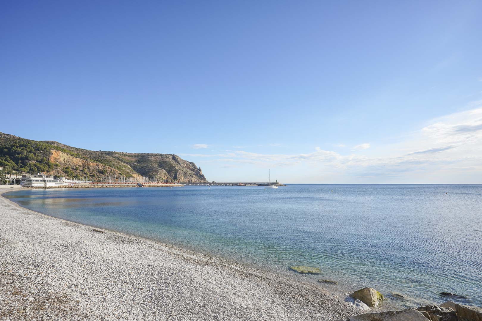Vista de la playa de grava en Jávea, con aguas cristalinas y el puerto deportivo al pie de los acantilados del Cabo de San Antonio.
