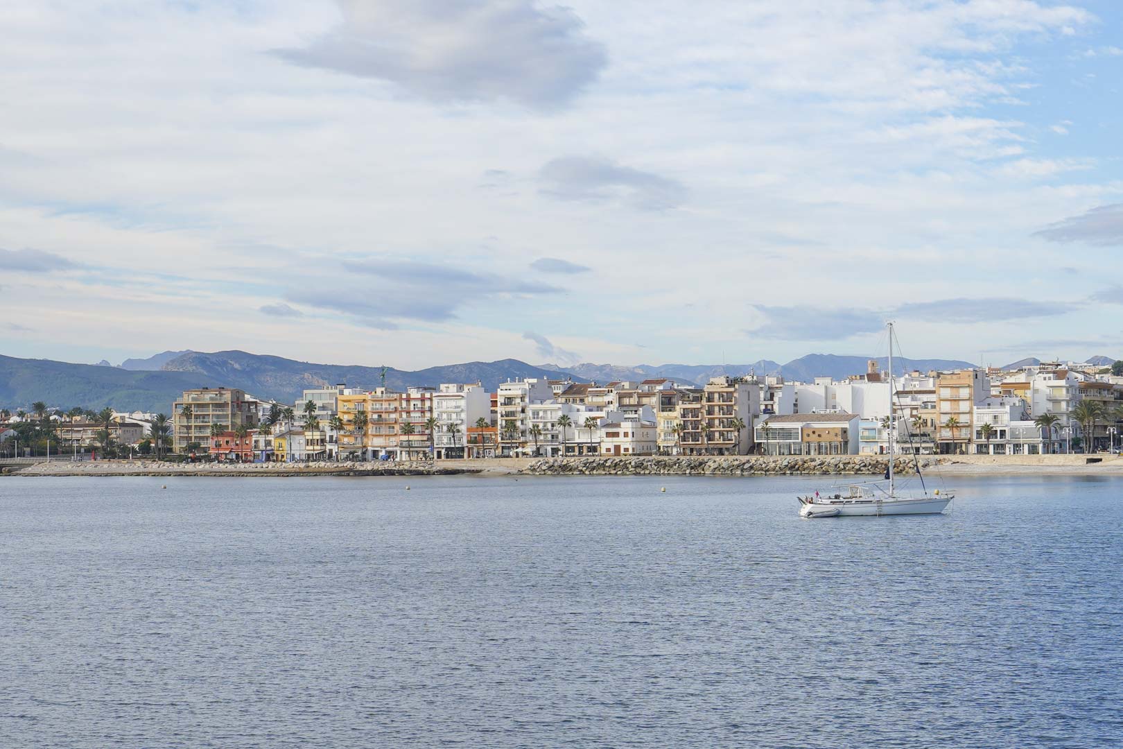 Vista panorámica del puerto de Jávea desde el mar, mostrando la arquitectura de primera línea, un velero en calma y las montañas al fondo bajo un cielo suave.
