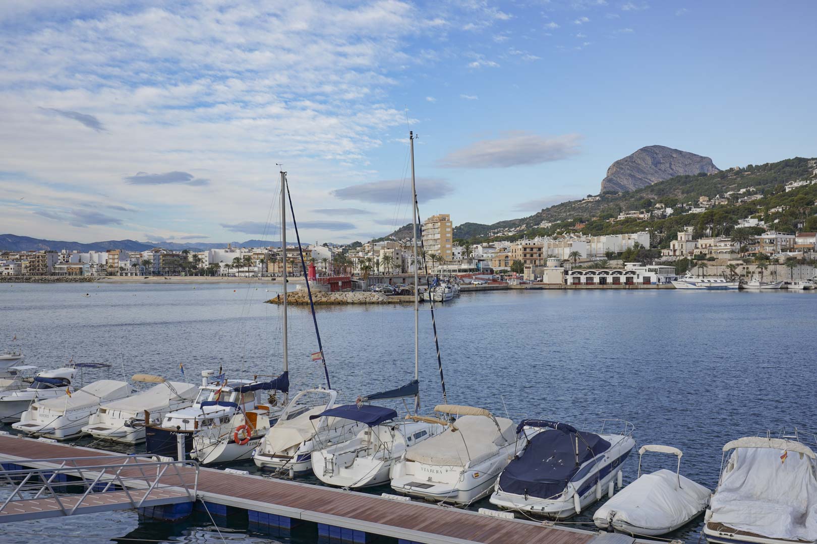 Vistas panorámicas del puerto de Jávea con embarcaciones de recreo amarradas y el perfil del macizo del Montgó bajo un cielo con nubes ligeras.