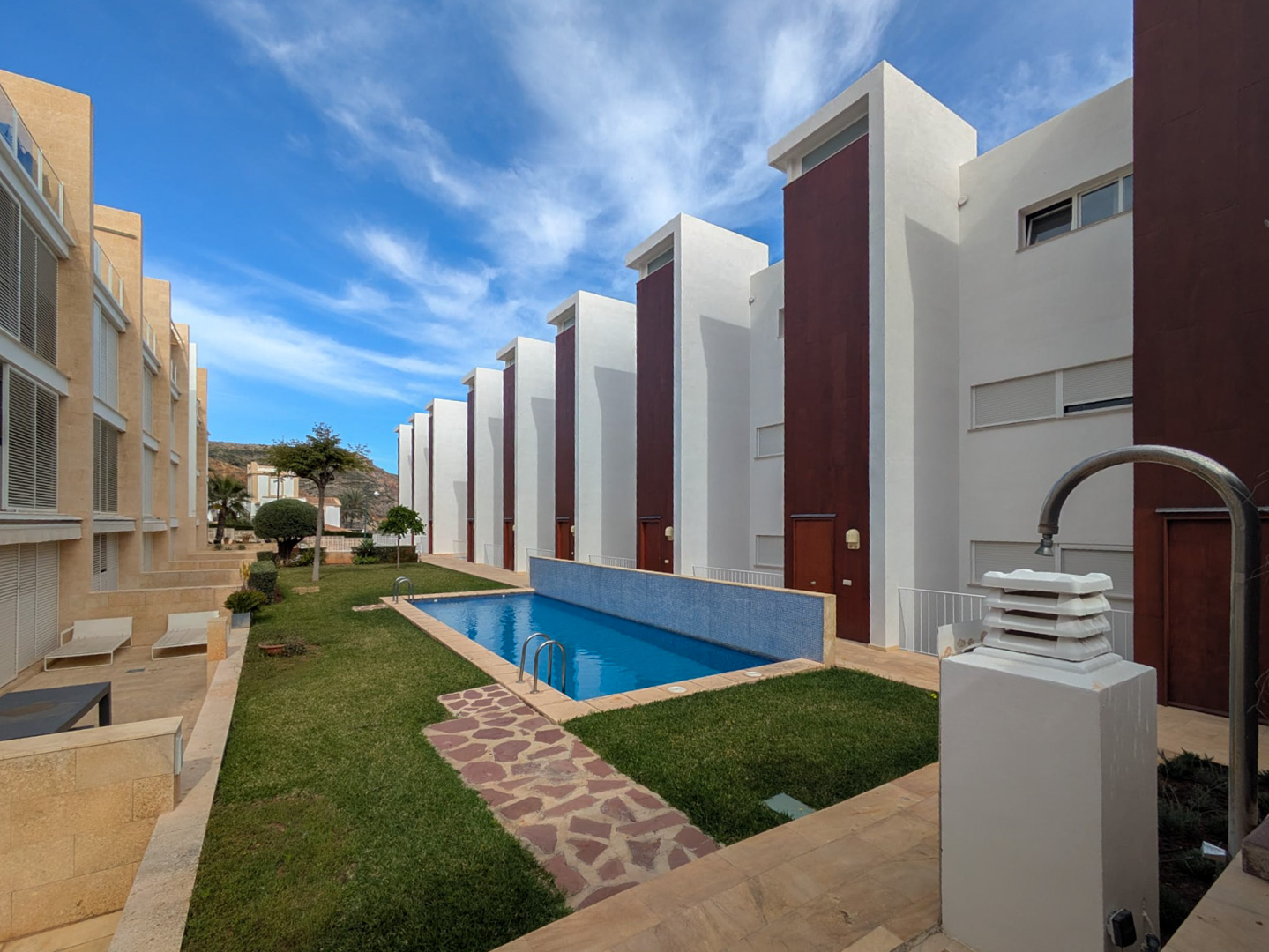 Zona comunitaria con piscina rectangular de gresite azul, rodeada de césped natural y senderos de piedra, integrada en un complejo de arquitectura moderna en Jávea.