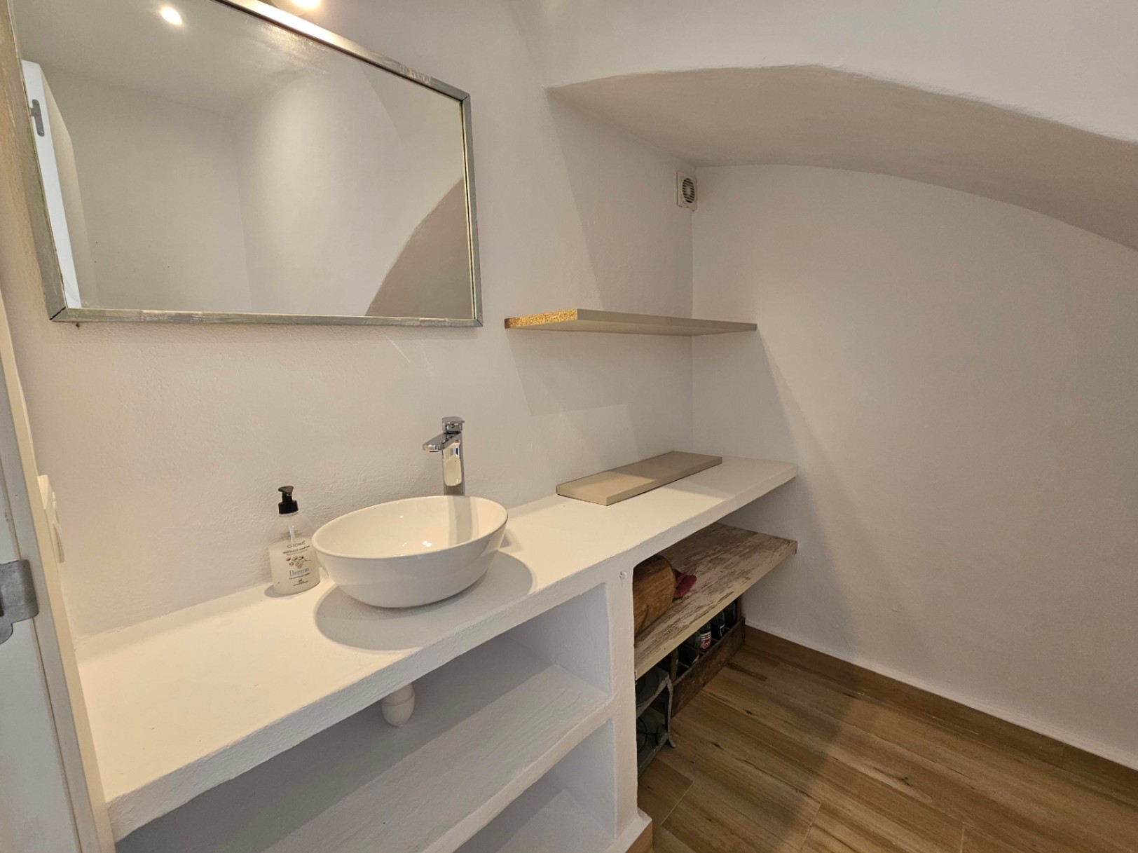 Aseo de cortesía con lavabo sobre encimera blanca de obra, estanterías integradas, suelo cerámico de efecto madera y techo abovedado bajo escalera.