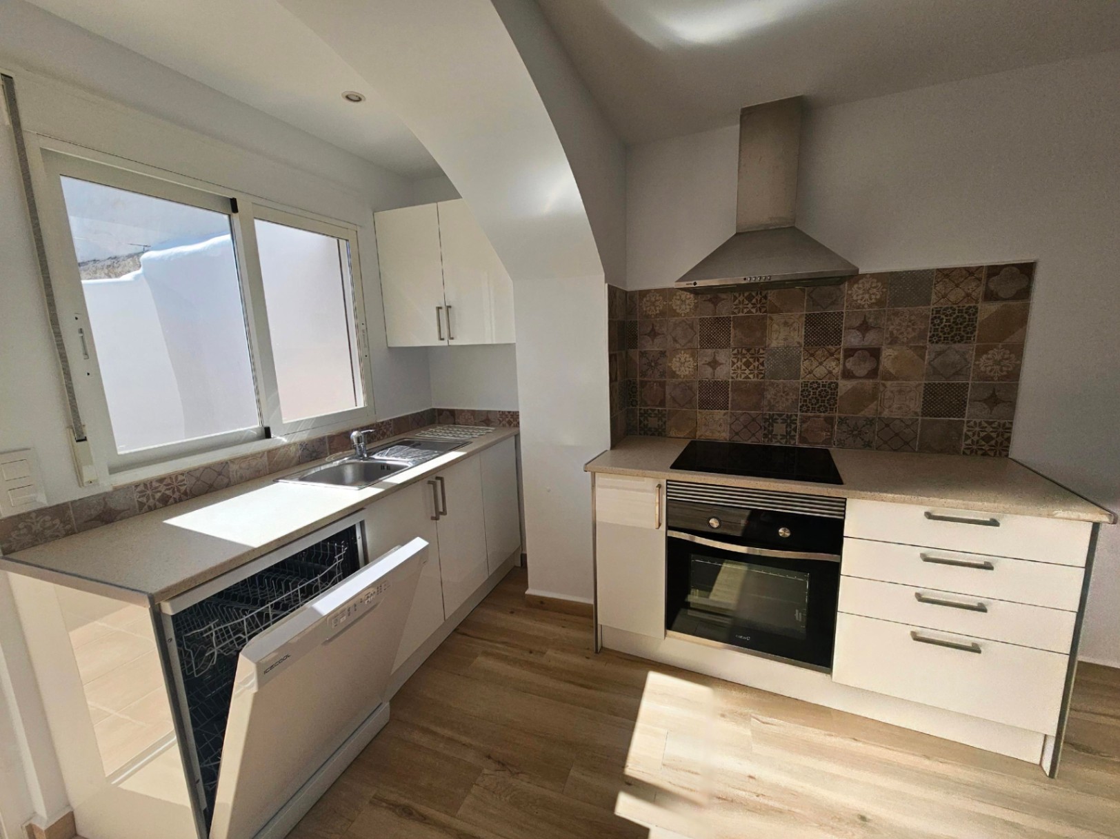 Cocina moderna con armarios blancos, encimera de piedra y salpicadero de azulejos hidráulicos. Incluye electrodomésticos integrados y luz natural del patio.