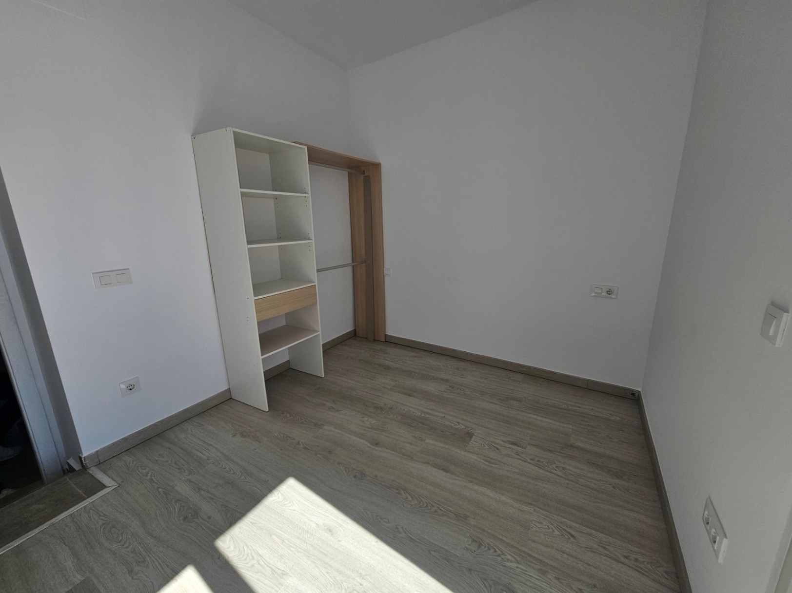 Dormitorio con suelos de tarima gris, paredes blancas y un armario abierto modular que combina estanterías blancas con detalles en madera clara.