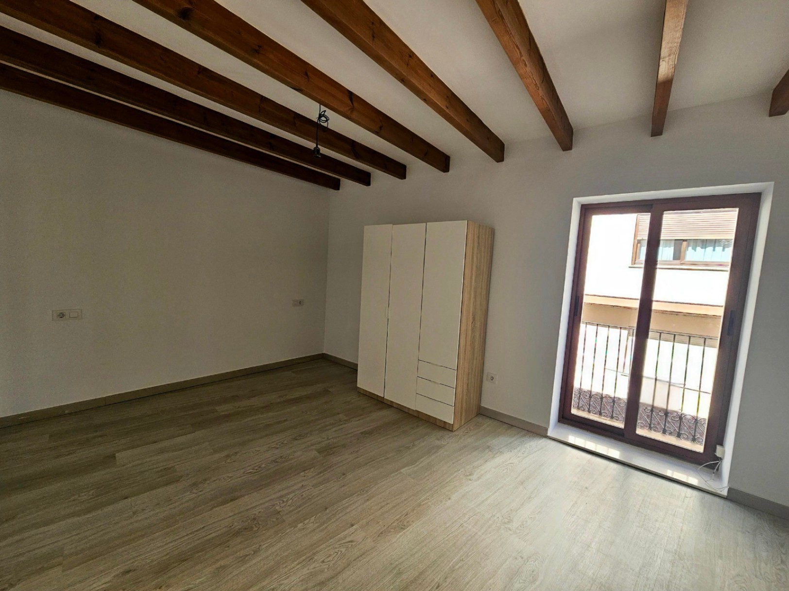 Dormitorio con vigas de madera vista, suelos de tarima clara y acceso directo a un balcón privado a través de puertas correderas de doble acristalamiento.