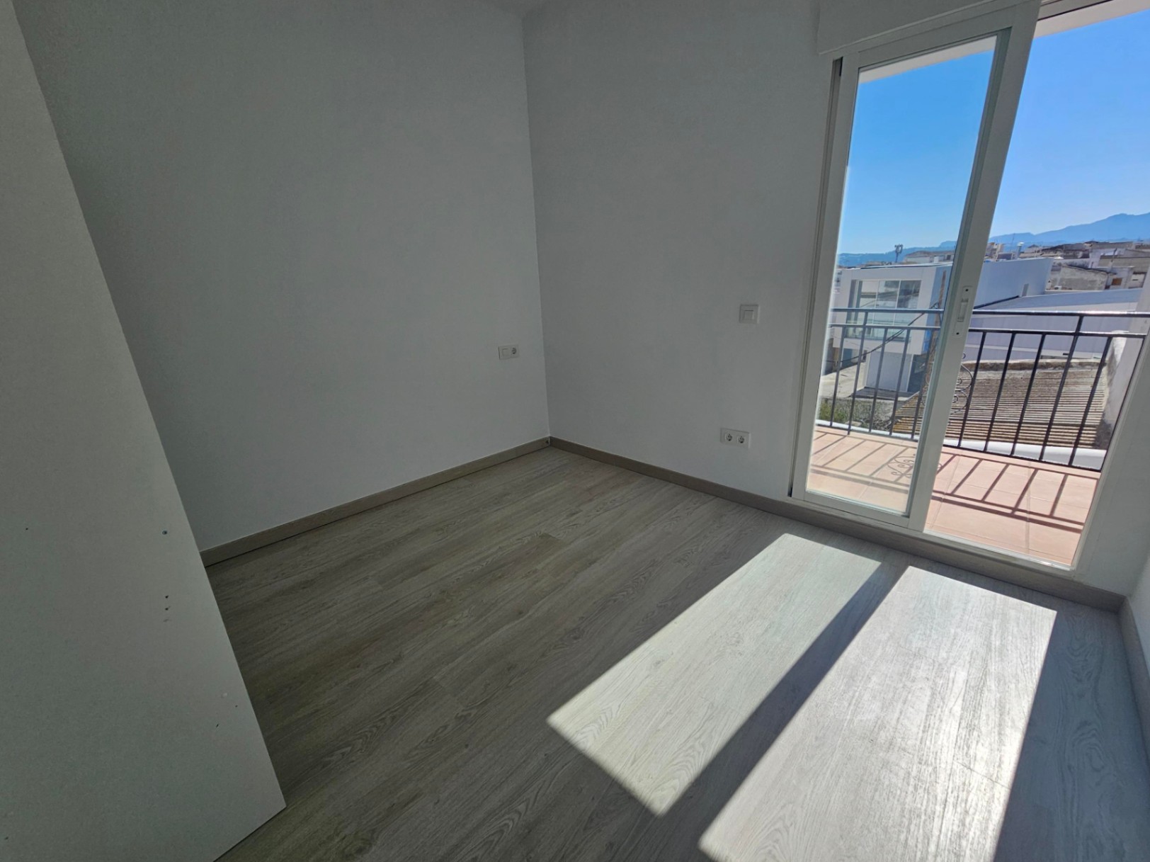 Dormitorio luminoso con suelos de madera clara y acceso directo a un balcón privado con vistas despejadas al pueblo y las montañas.