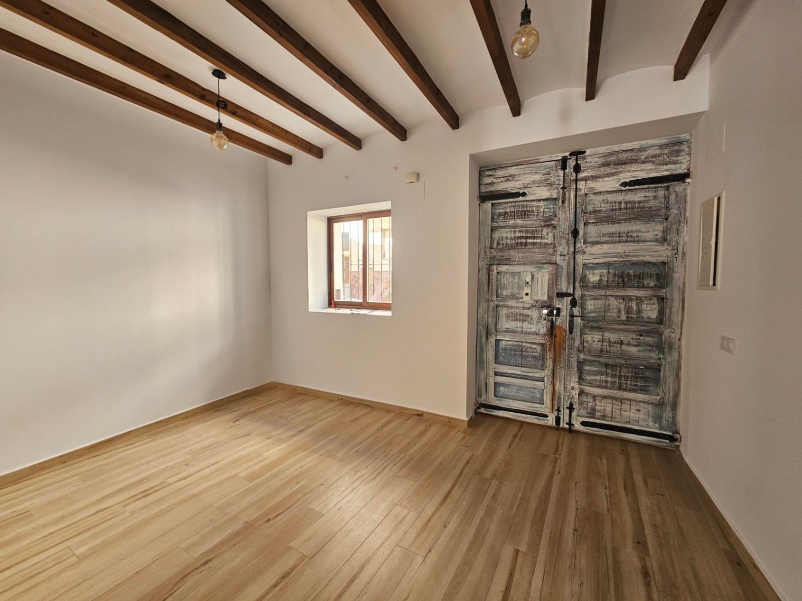 Estancia con vigas de madera expuestas, suelos claros y una gran puerta rústica restaurada con herrajes de hierro. Incluye iluminación minimalista y ventana con marco de madera.