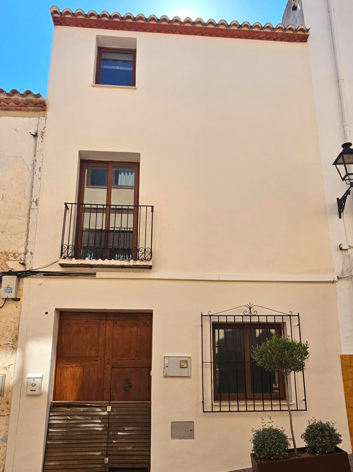 Fachada de tres niveles con paredes en tono crema, puerta principal de madera, balcón de forja negra y ventanas con marcos de madera y doble acristalamiento.