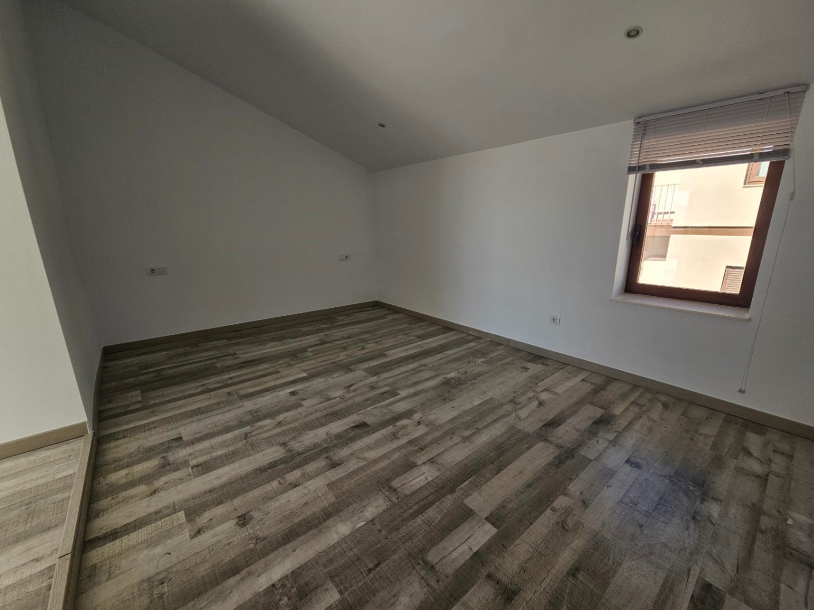 Habitación con techo abuhardillado, suelos laminados de efecto madera y focos empotrados. Dispone de ventana con marco de madera y persiana para luz natural.
