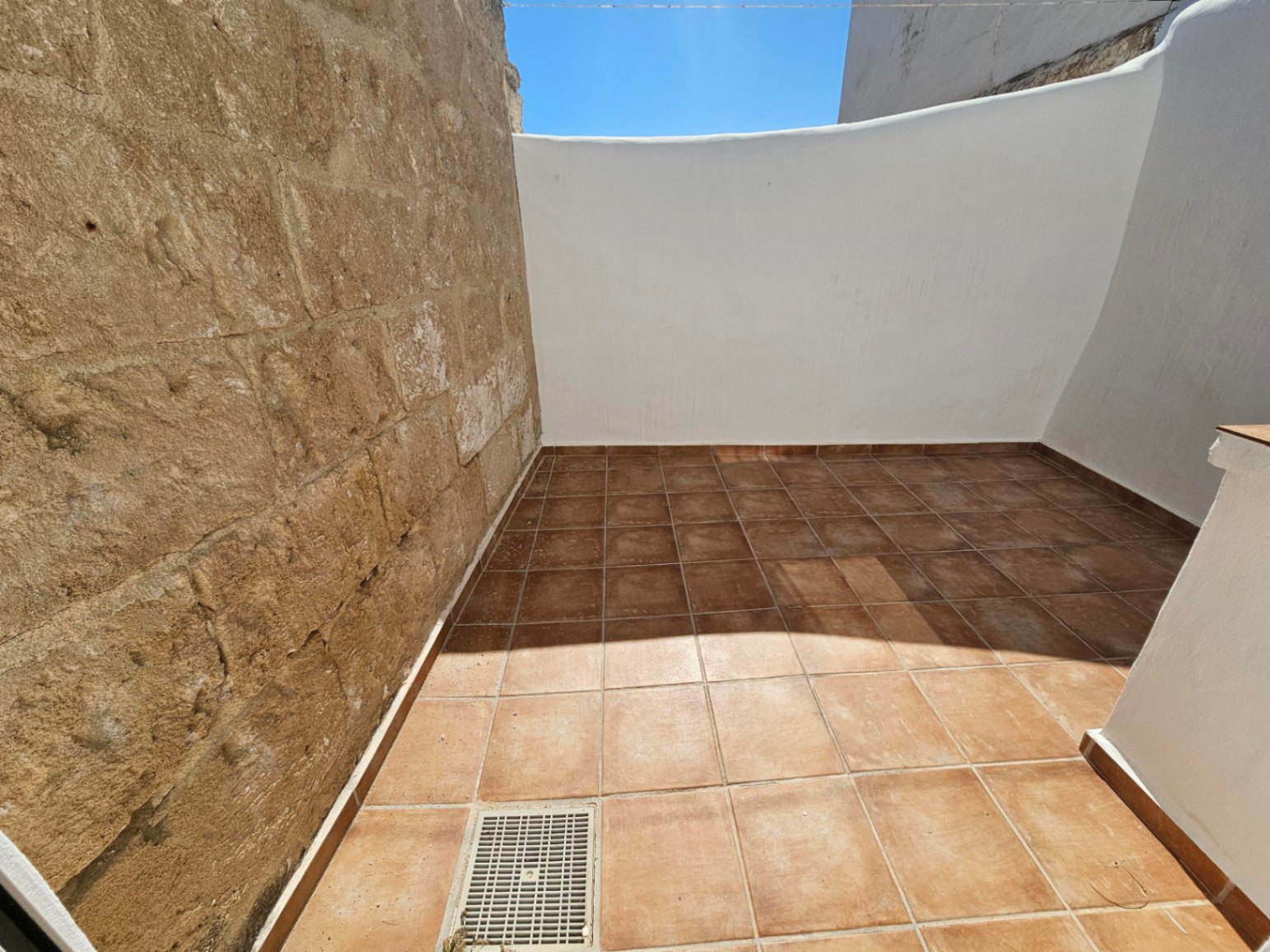 Patio exterior con suelo de baldosas de terracota, combinando una pared de piedra natural vista con muros blancos bajo el cielo despejado.