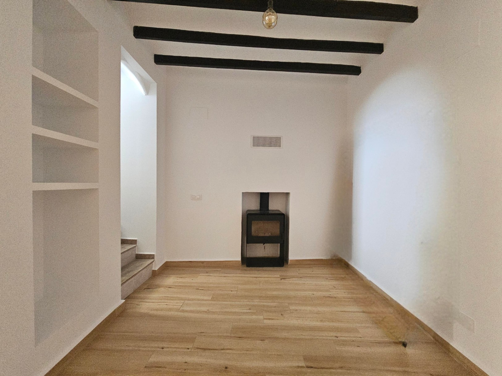 Salón con vigas de madera oscura, estantería de obra y quemador de leña encastrado. El suelo de tarima clara y las paredes blancas aportan luminosidad al espacio.