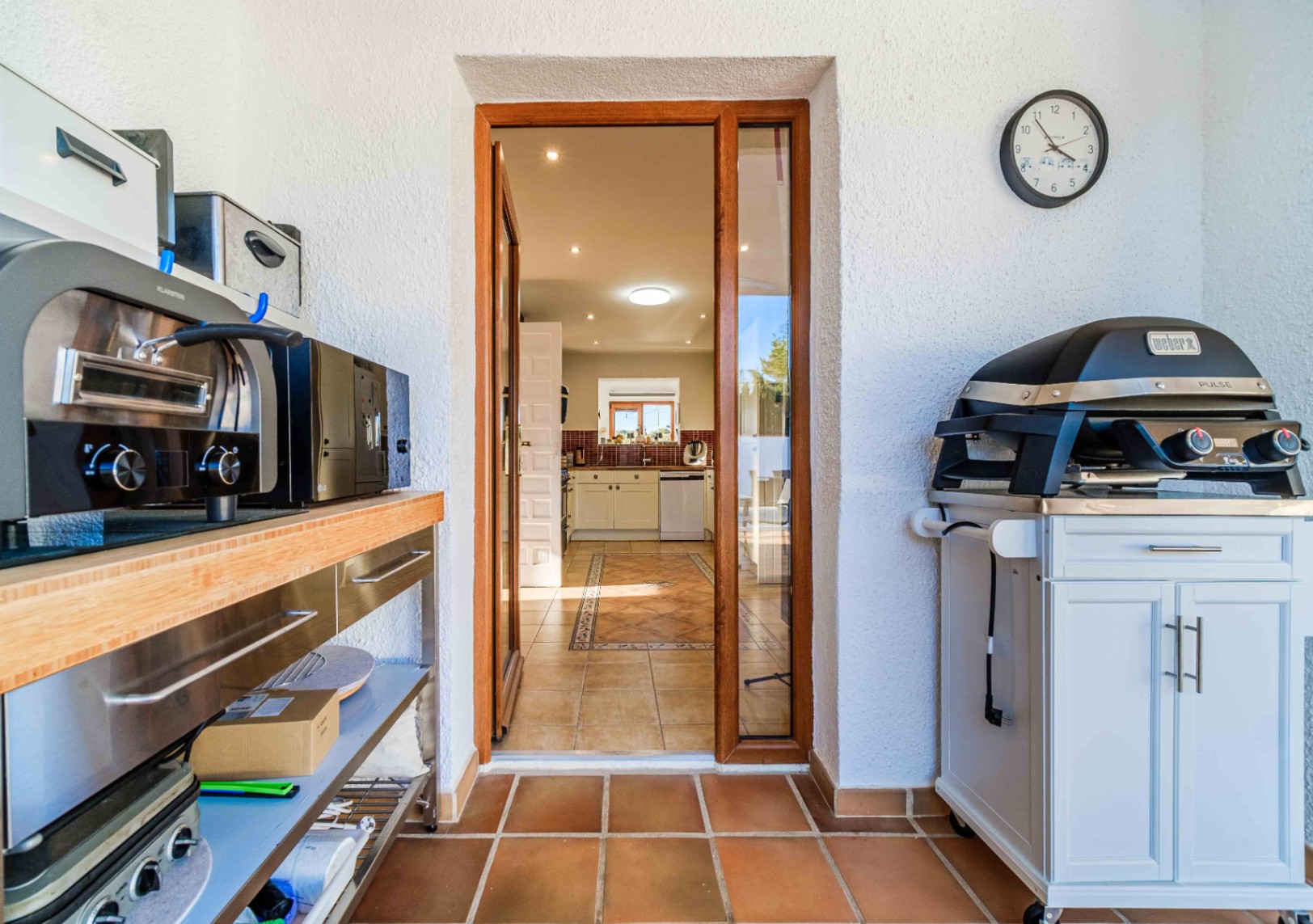 Área de cocina exterior equipada con barbacoa eléctrica Weber y encimera de madera, con acceso directo a la cocina principal de estilo rústico a través de una puerta acristalada.