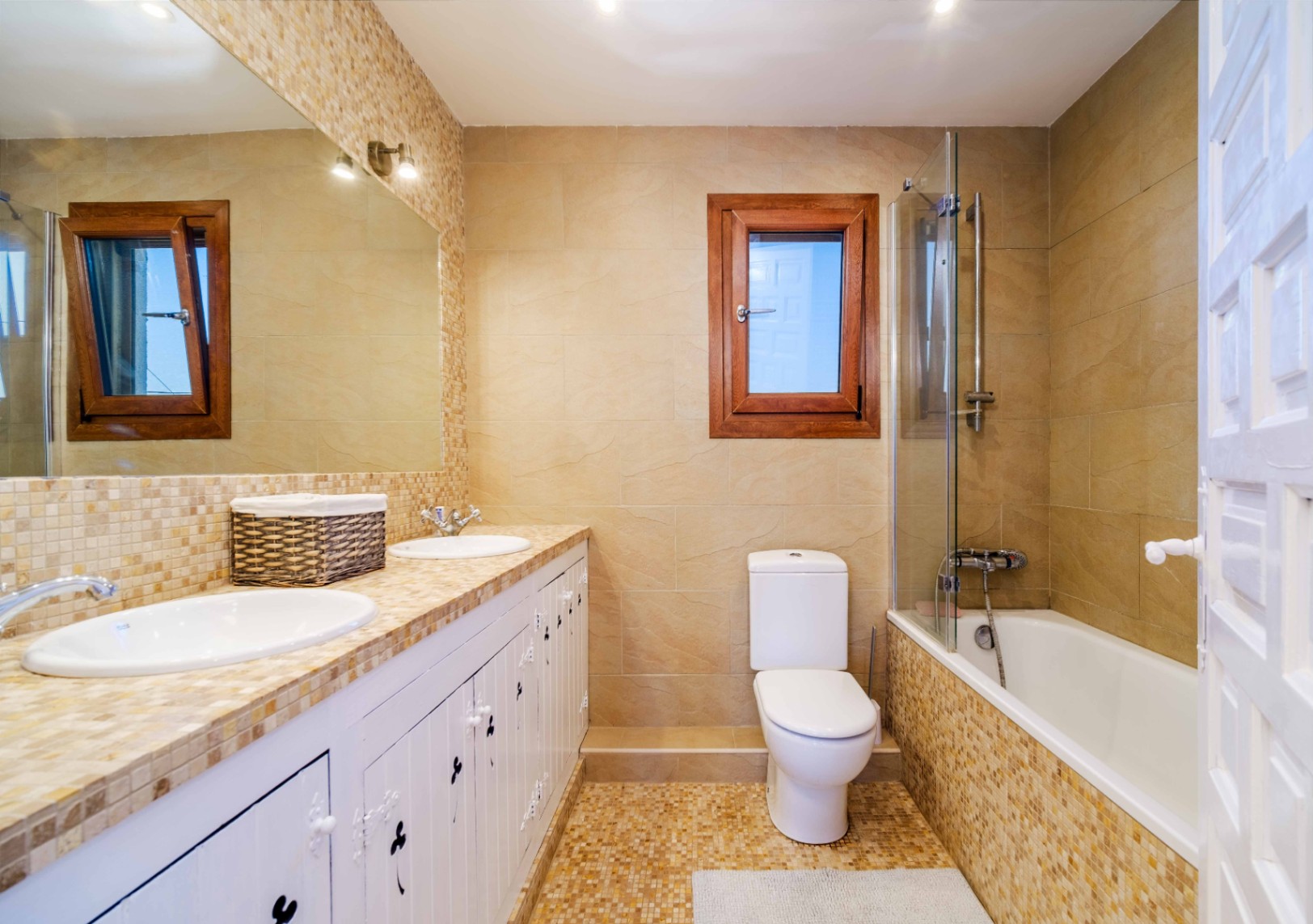 Baño completo con lavabo doble integrado en encimera de mosaico, mueble blanco de estilo rústico y bañera con mampara de cristal y revestimiento cerámico en tonos tierra.