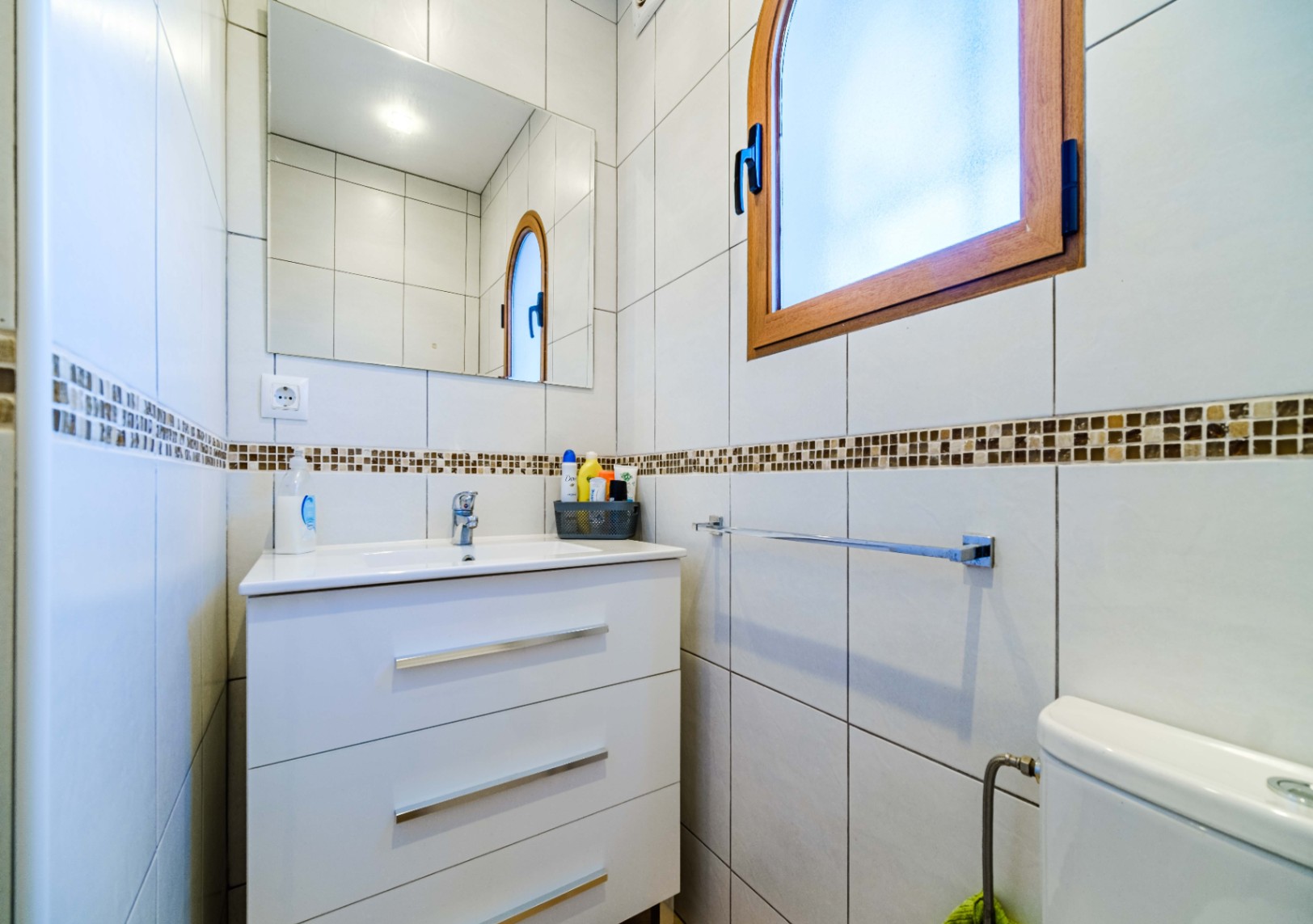 Baño funcional con mueble de lavabo blanco de tres cajones, espejo rectangular y ventana de madera en arco. Paredes alicatadas con cenefa de mosaico en tonos tierra.