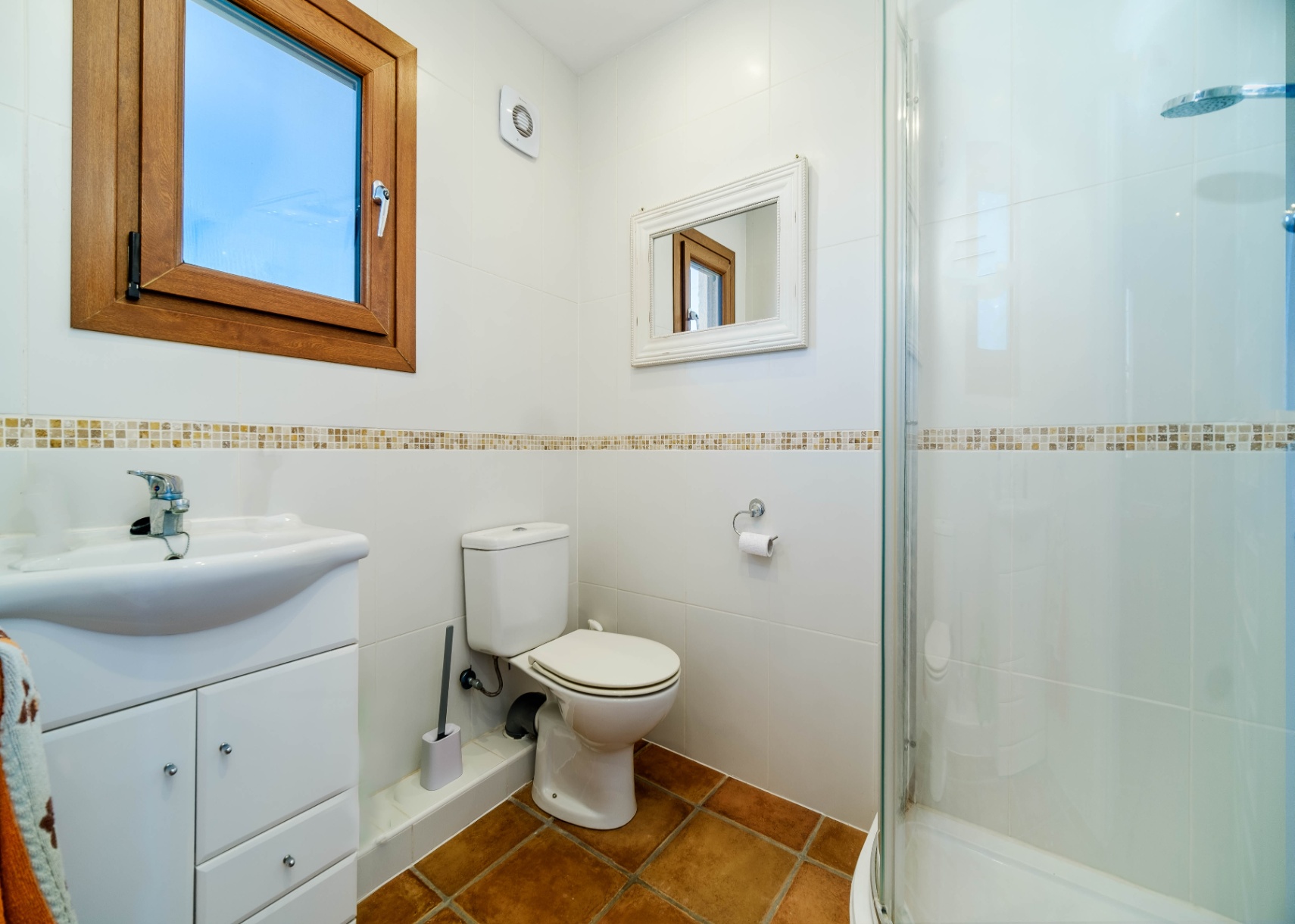 Baño funcional con paredes alicatadas en blanco, cenefa decorativa, suelo de terracota y ducha con mampara de cristal.