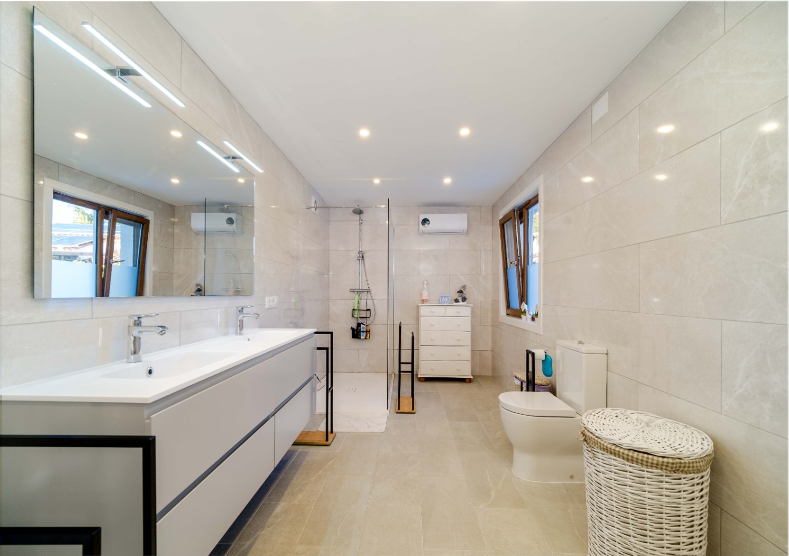 Baño moderno con lavabo doble, mueble suspendido gris y gran espejo iluminado. Incluye ducha a ras de suelo con mampara de cristal y revestimiento cerámico en tonos neutros.
