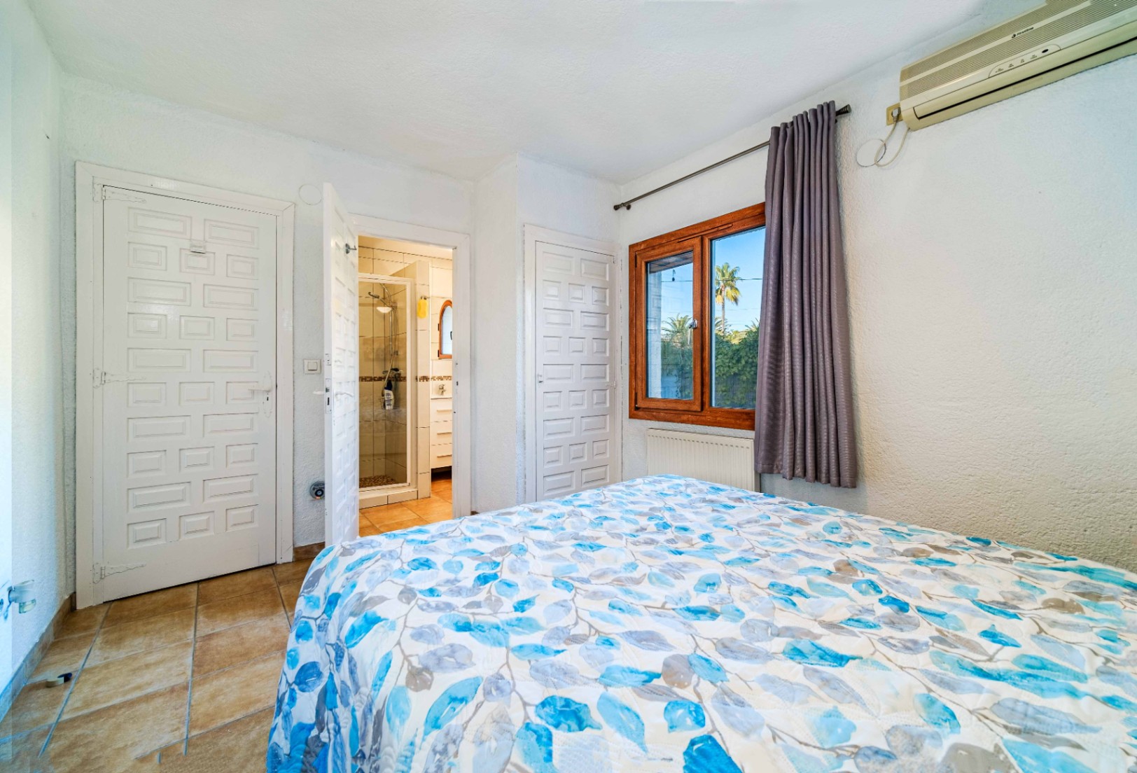 Dormitorio con baño en suite, puertas blancas de cuarterones, suelos cerámicos y ventana de madera. Equipado con aire acondicionado y calefacción por radiador.