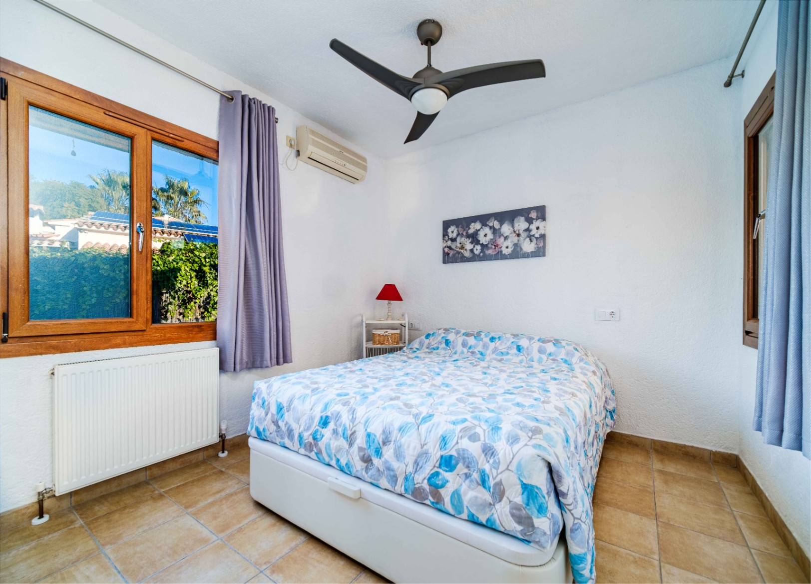 Dormitorio luminoso con suelos de gres, ventilador de techo, aire acondicionado y ventanas de madera que ofrecen vistas al jardín mediterráneo.