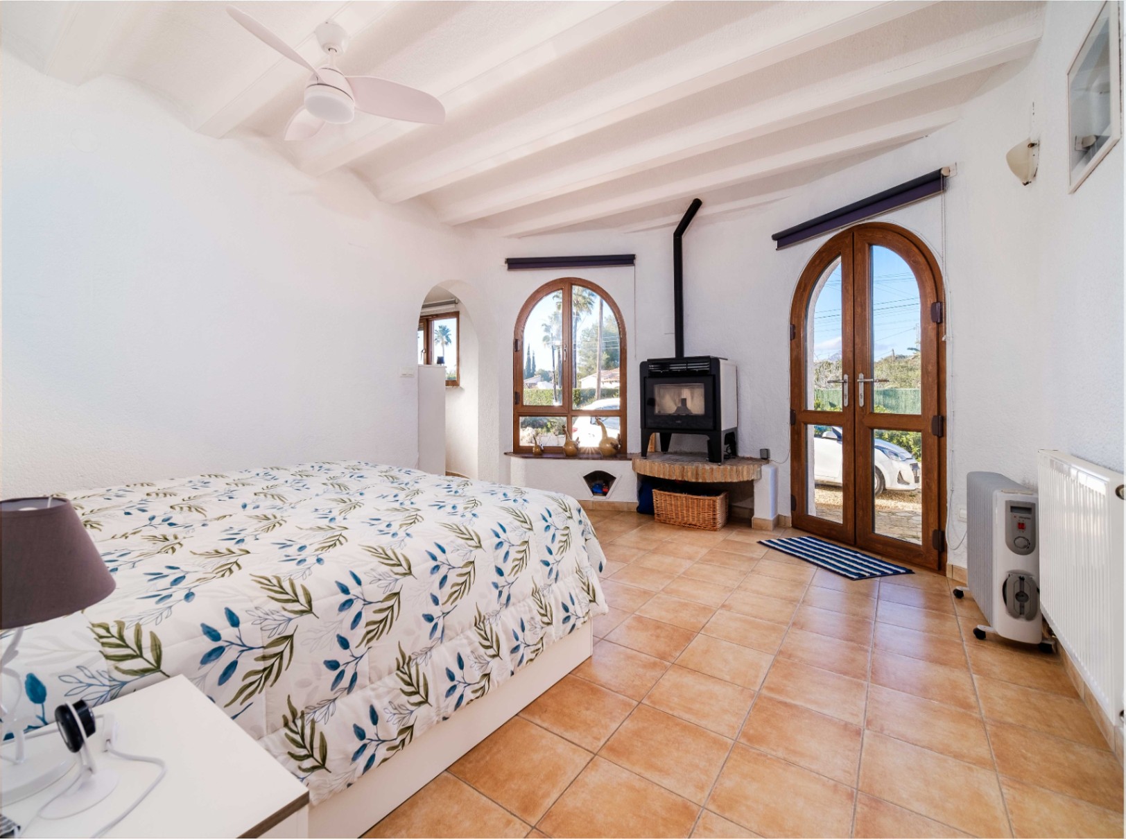 Dormitorio luminoso con techos de vigas blancas, ventilador y estufa de leña. Cuenta con suelos de terracota y acceso directo al exterior mediante puertas de madera en arco.