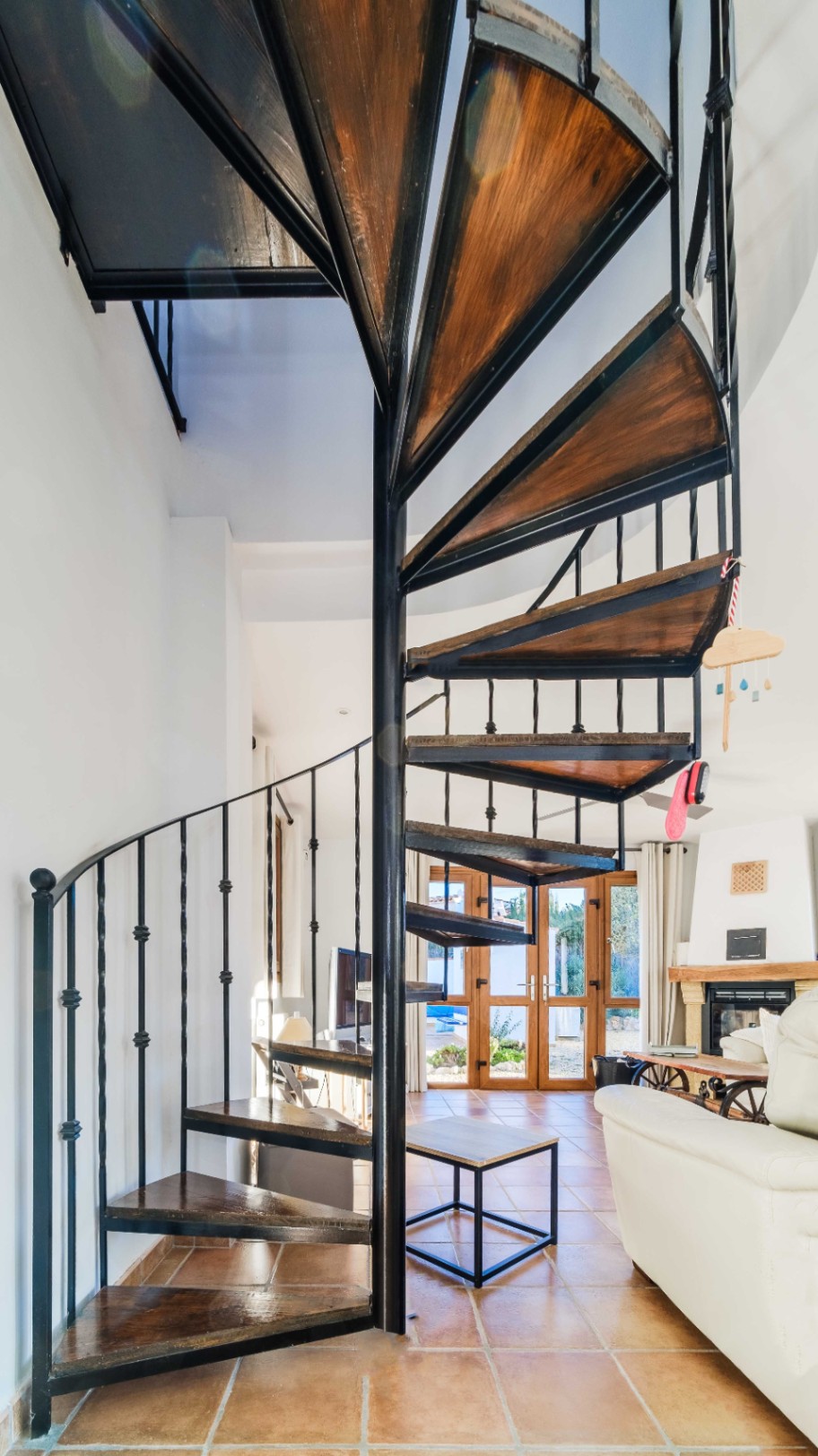 Escalera de caracol con estructura de hierro forjado negro y peldaños de madera oscura, integrada en un salón con suelos de terracota y chimenea.