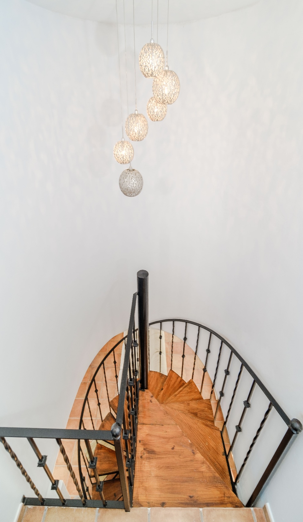 Escalera de caracol con peldaños de madera y barandilla de hierro forjado, iluminada por un conjunto de lámparas colgantes esféricas sobre paredes blancas.