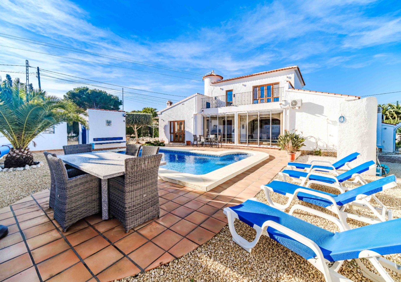 Exterior de la villa con piscina, terraza de baldosas de barro, zona de comedor exterior y tumbonas azules frente a la fachada blanca mediterránea.