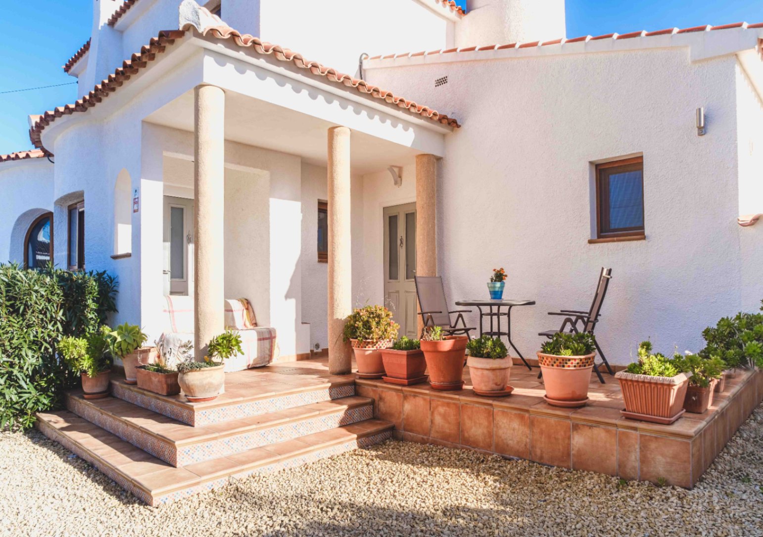 Fachada blanca con porche sostenido por columnas de piedra, suelos de terracota y escalones con detalles de mosaico. Incluye una zona de estar exterior decorada con plantas.