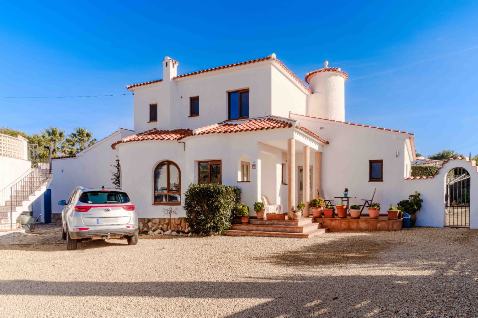 Fachada mediterránea de estuco blanco con tejado de teja cerámica, porche de columnas y amplia zona de aparcamiento de grava bajo un cielo despejado.