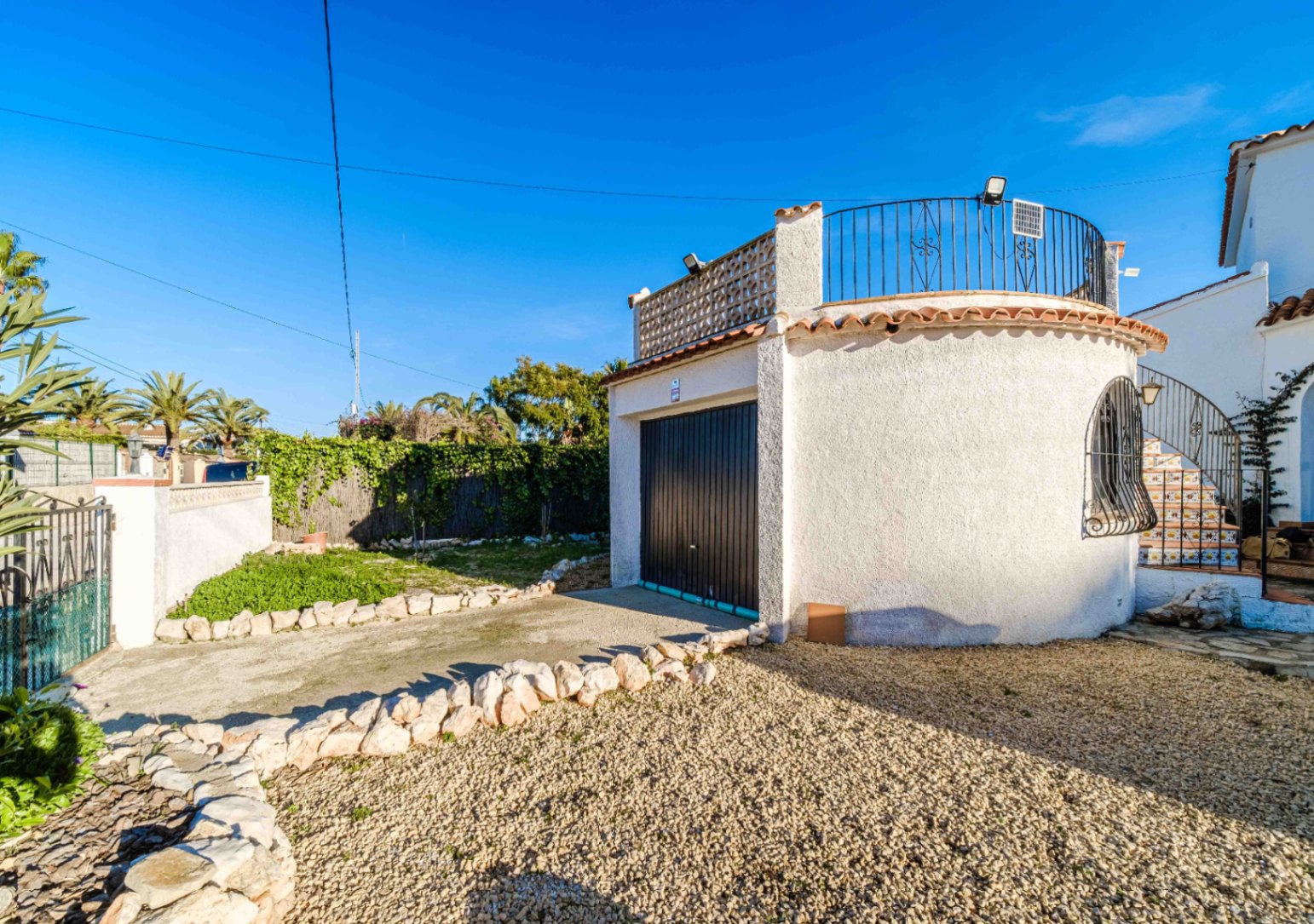 Garaje privado con puerta negra y terraza superior accesible mediante escaleras decoradas. El entorno incluye grava, bordes de piedra natural y vegetación mediterránea.