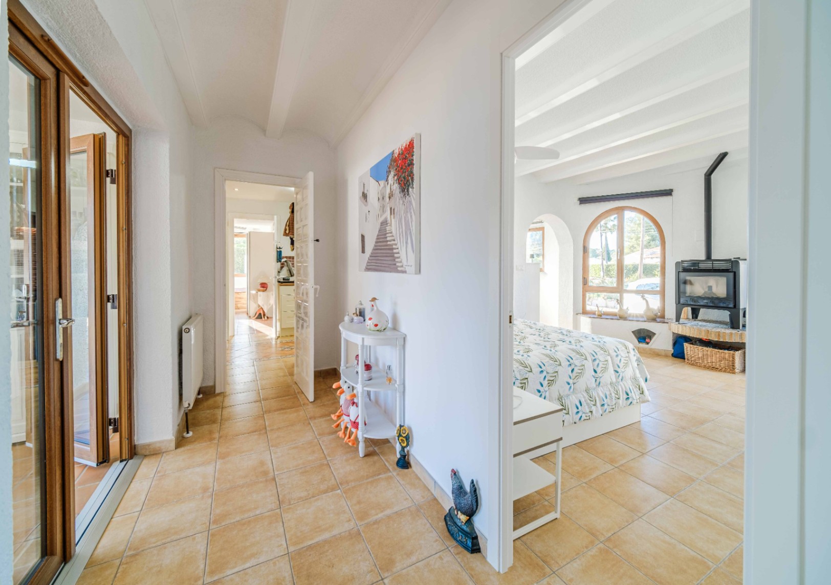 Pasillo con suelos cerámicos que conecta estancias, destacando un dormitorio con techos de vigas blancas, ventana de arco y estufa de leña independiente.