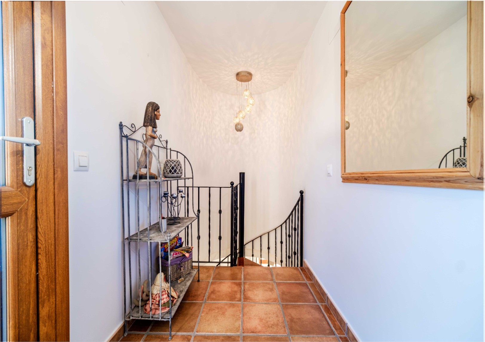 Pasillo distribuidor con suelos de terracota, barandilla de forja negra y una escalera curva iluminada por una lámpara colgante decorativa.
