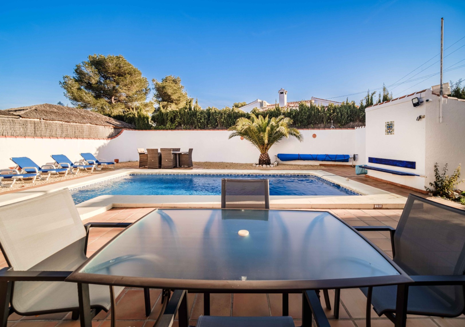 Piscina rectangular rodeada de una terraza de gres, zona de tumbonas sobre grava y comedor exterior. El espacio cuenta con muros perimetrales blancos y vegetación mediterránea.