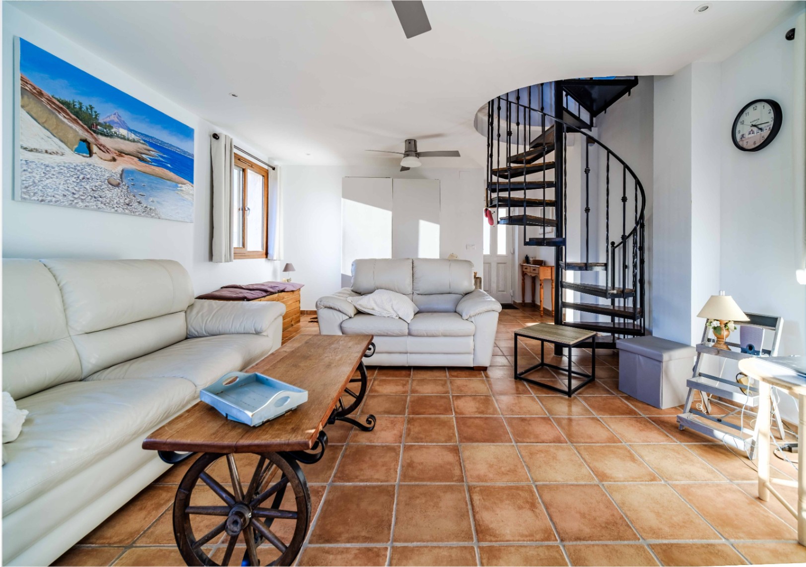 Salón con suelos de terracota, sofás de cuero blanco y una distintiva escalera de caracol metálica que conecta con el nivel superior.
