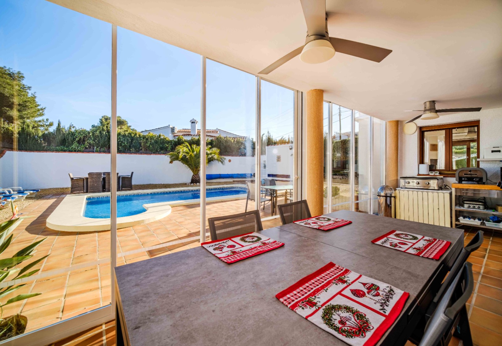 Terraza acristalada con comedor, ventiladores de techo y cocina exterior, ofreciendo vistas despejadas a la piscina de agua salada y al jardín.