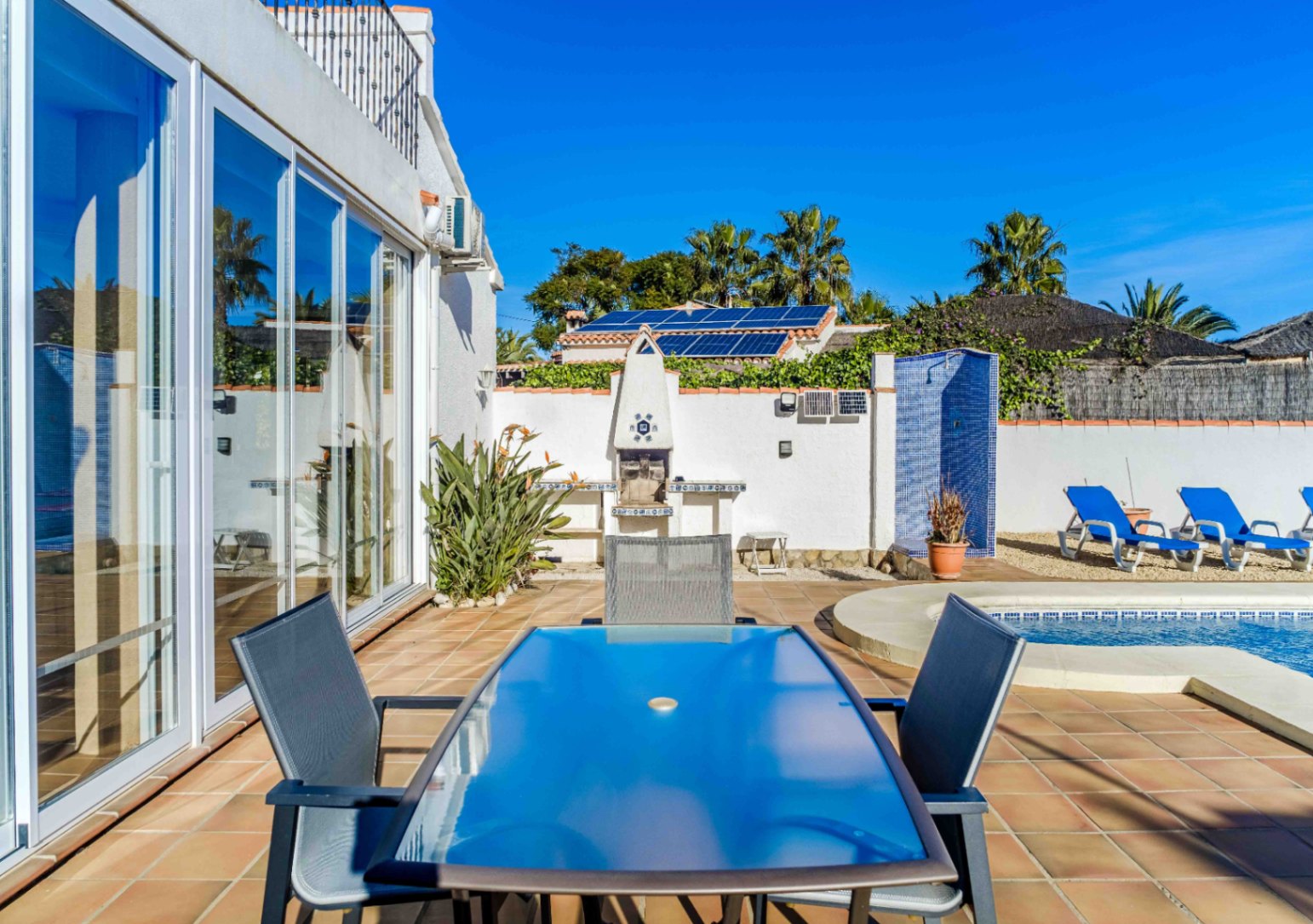 Terraza exterior con zona de comedor, barbacoa integrada y ducha de gresite azul junto a la piscina. Incluye amplios ventanales correderos y paneles solares visibles.